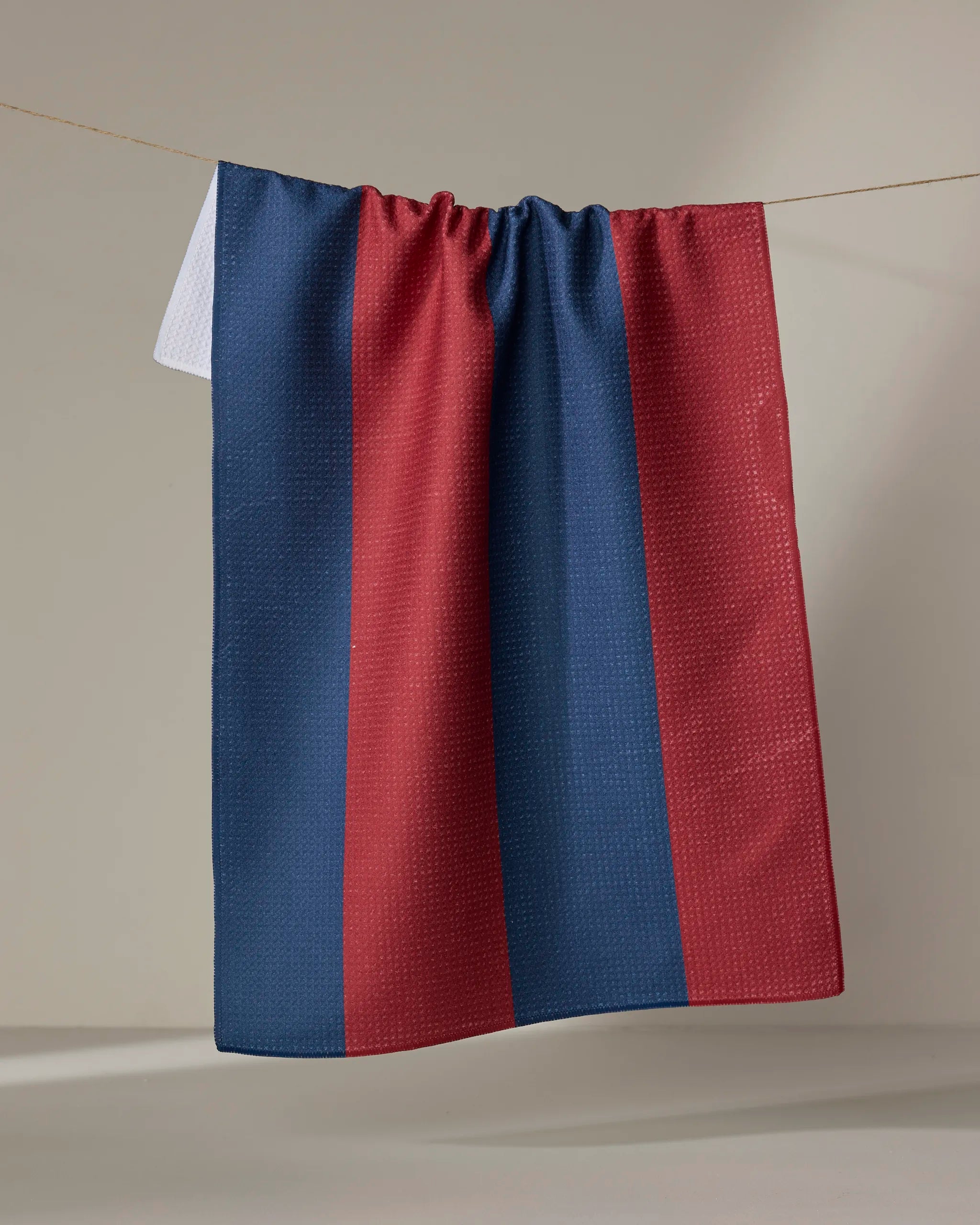 The Modern Dad’s Mr. Rugby tea towel, featuring quick-drying fabric and vertical blue and maroon stripes, hangs on a clothesline against a plain light background.
