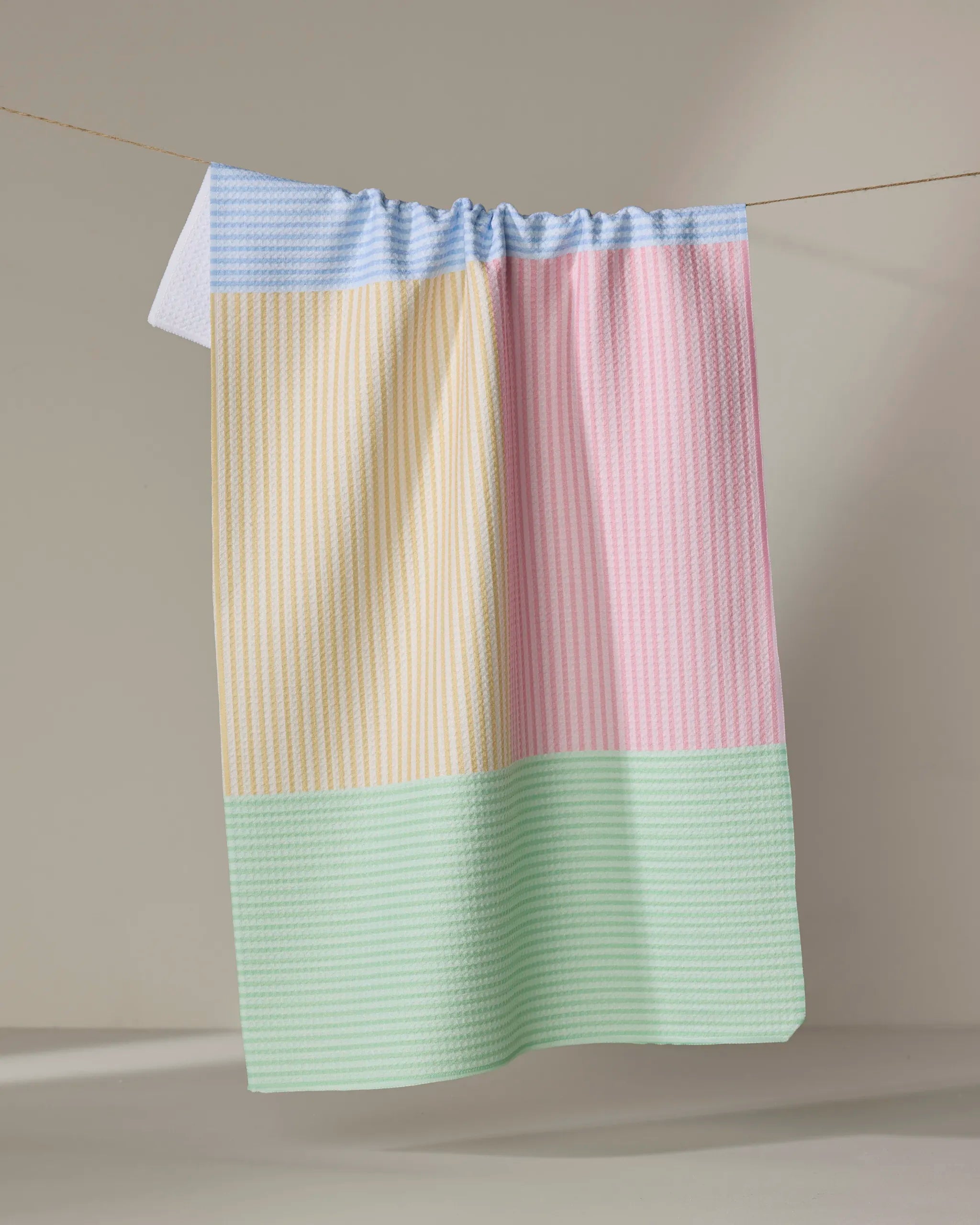 The Modern Dad’s Mr. Fun Stripes kitchen tea towel features a pastel patchwork in blue, yellow, pink, and green. Ultra-absorbent and stylish, it hangs on a line against a plain beige background—practical for daily use.
