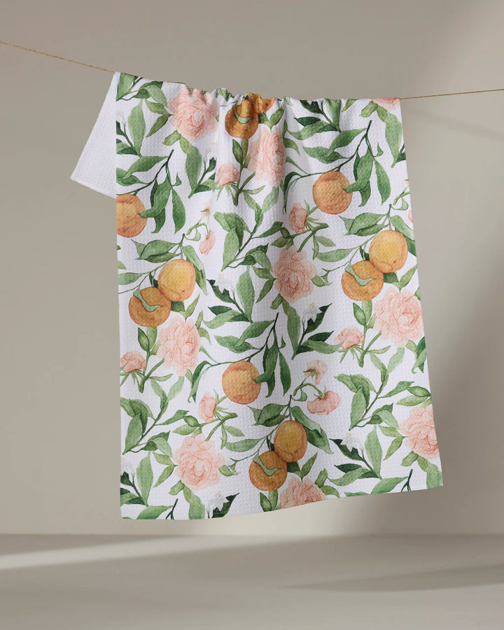 The "A Peony For Your Thoughts" recycled kitchen towel by Aubrey Fairchild, with a vibrant floral and foliage pattern, hangs on a line against a plain beige background.