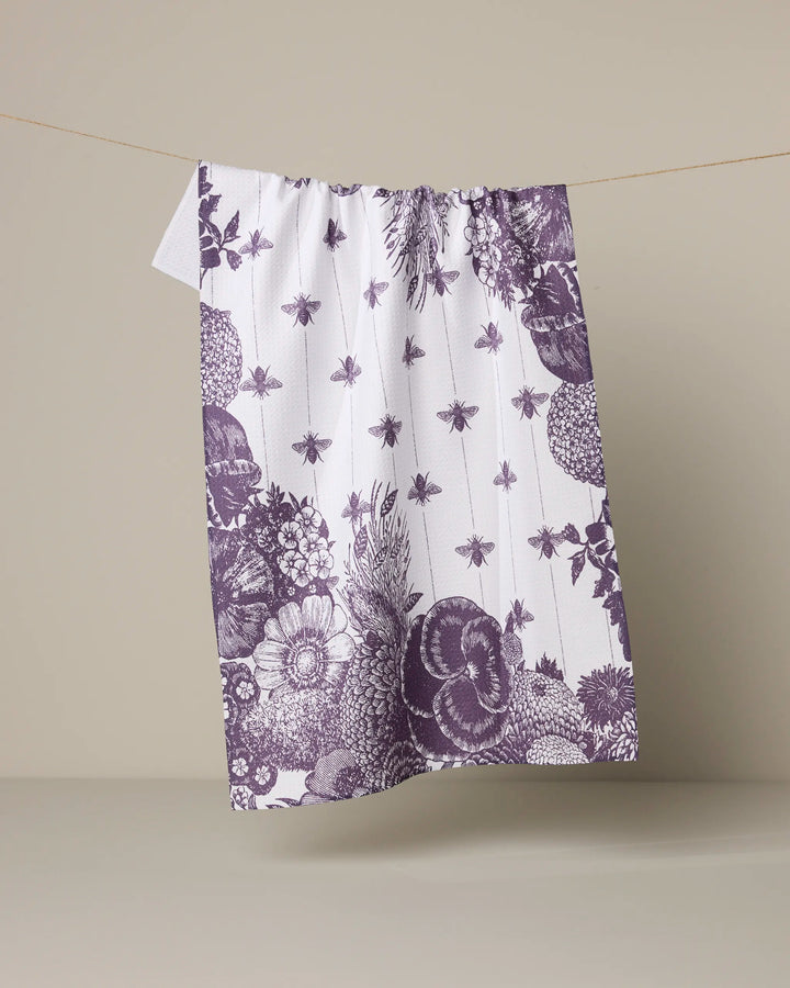 The Abeilles Noires tea towel by Geometry features a detailed vintage pattern of purple botanicals, fruits, and bees on white, and is shown hanging against a plain beige background.