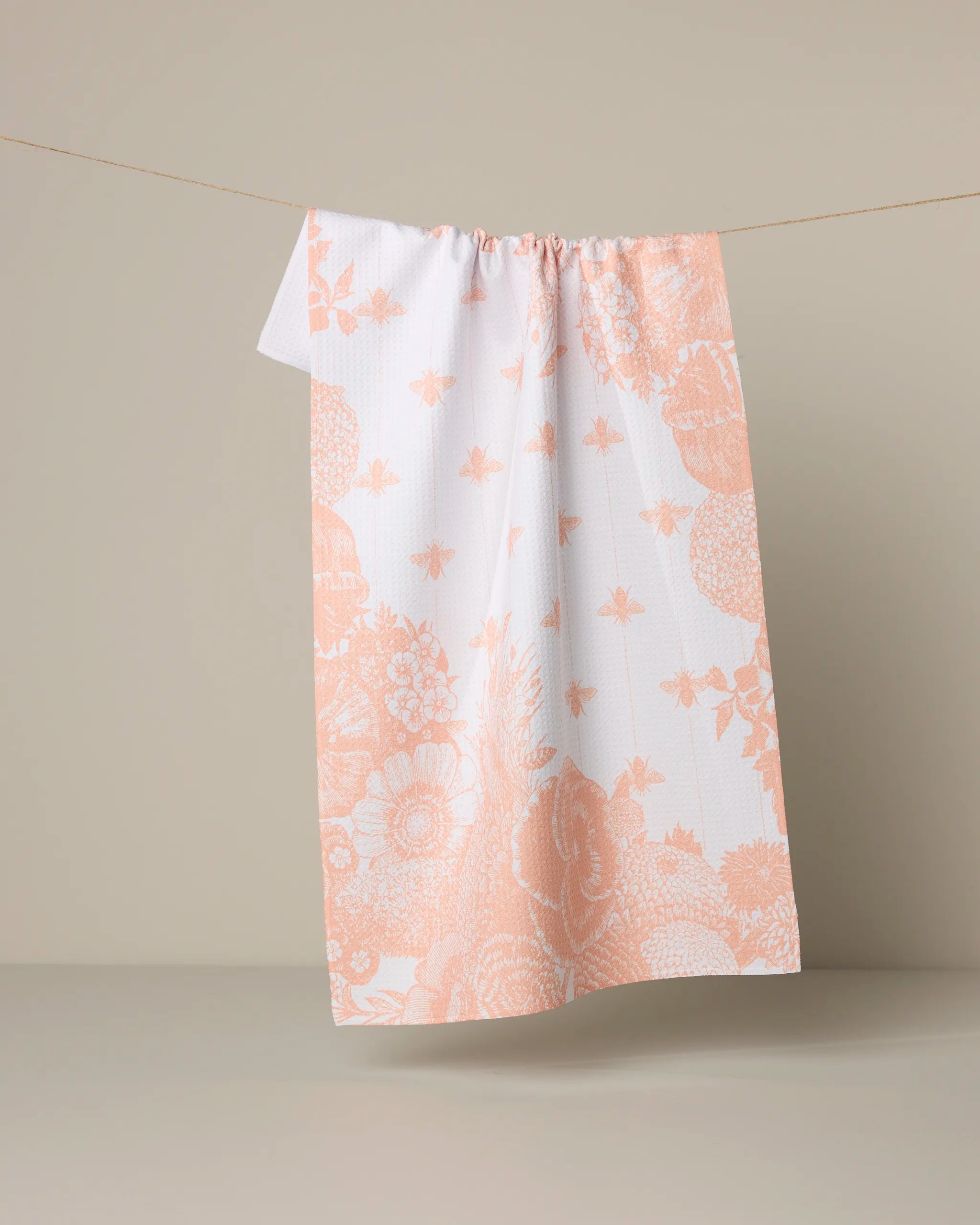 The Geometry Abeilles kitchen towel features a peach floral and leaf design on white and hangs from a string against a plain beige backdrop.
