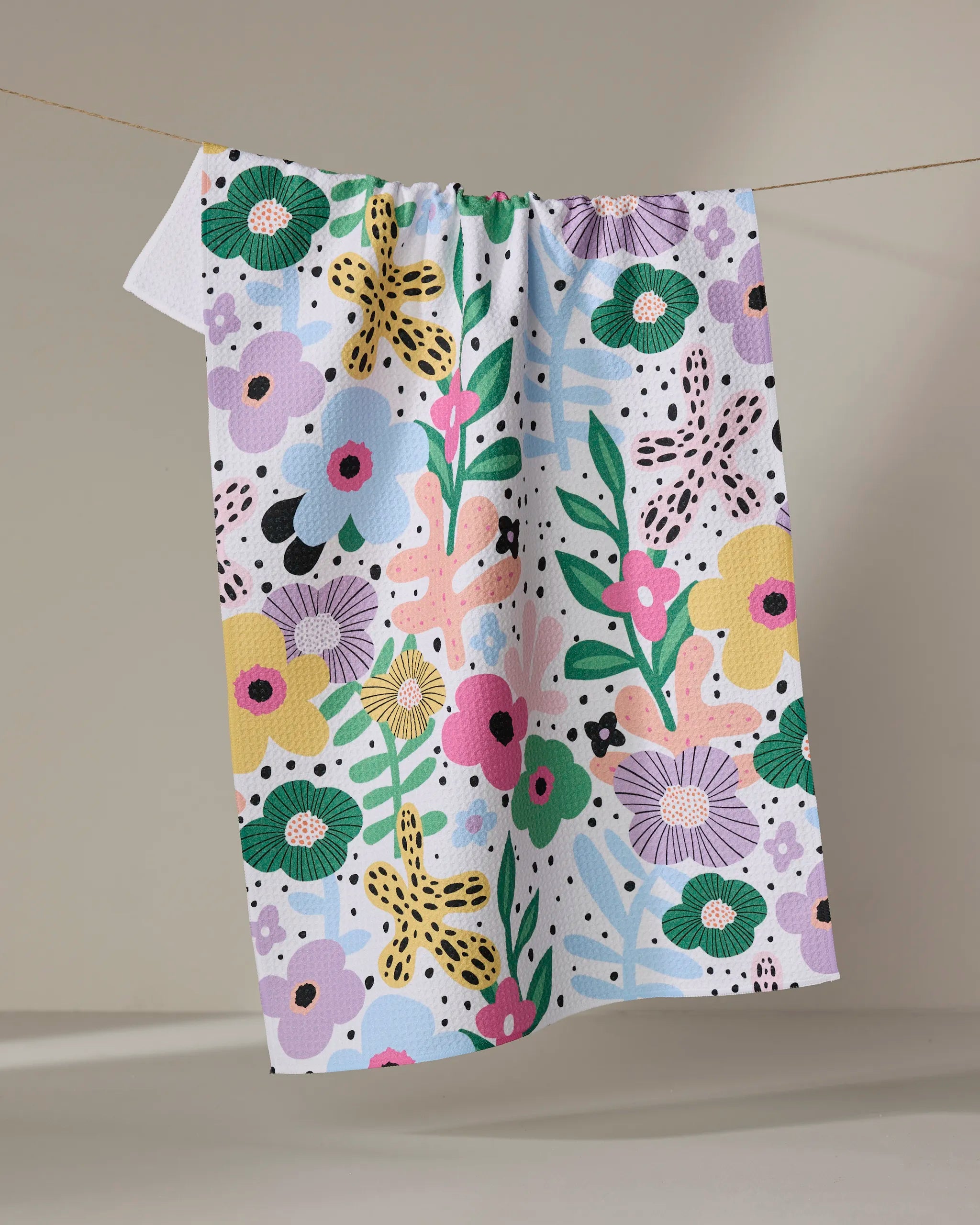 The "Abstract Botanicals" tea towel by Marina Solodka, made from recycled materials and featuring a vivid floral and leaf print in green, yellow, pink, purple, and blue, hangs on a string against a neutral background.