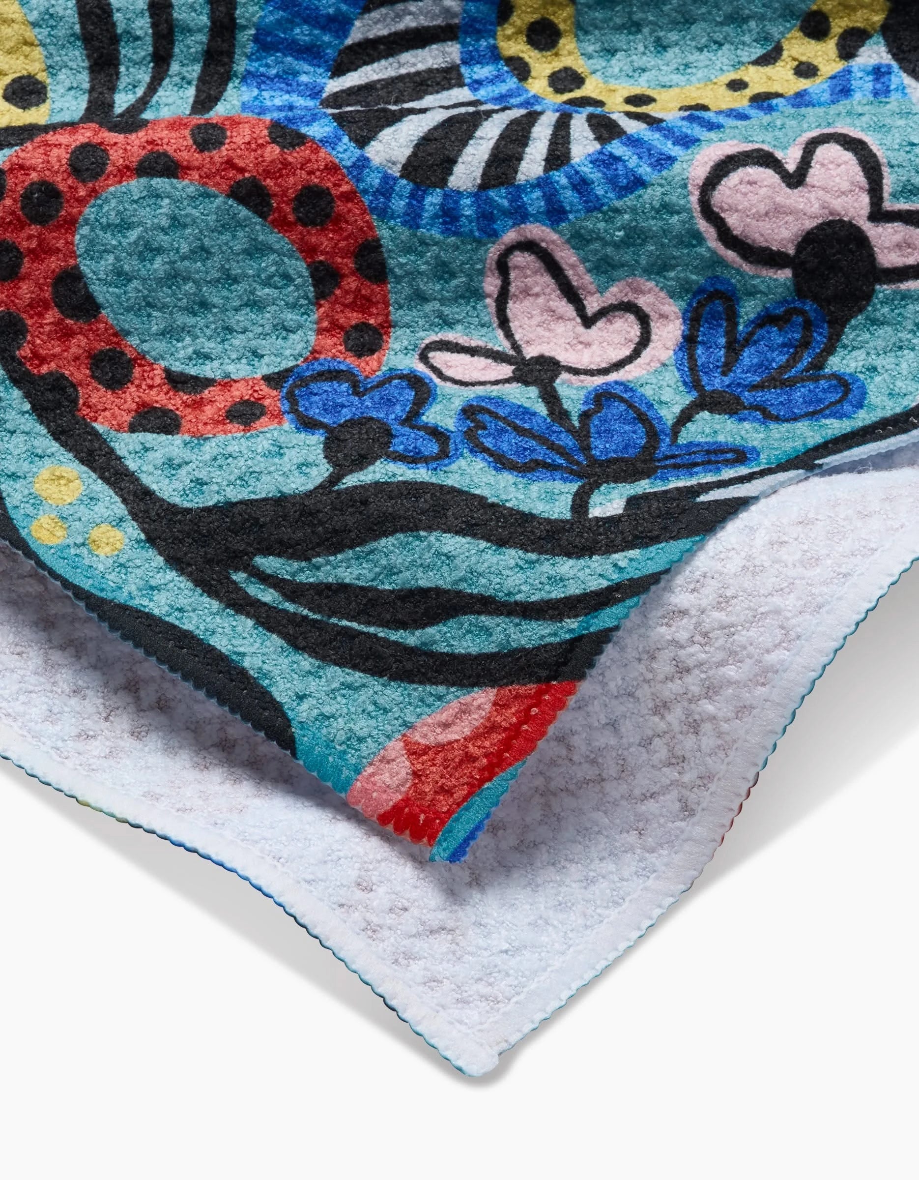 A close-up of the Abstract Dreams tea towel by Masha Volnova showcases abstract shapes, flowers, and bold outlines on Geoweave fabric, with the white textured reverse side partially visible.