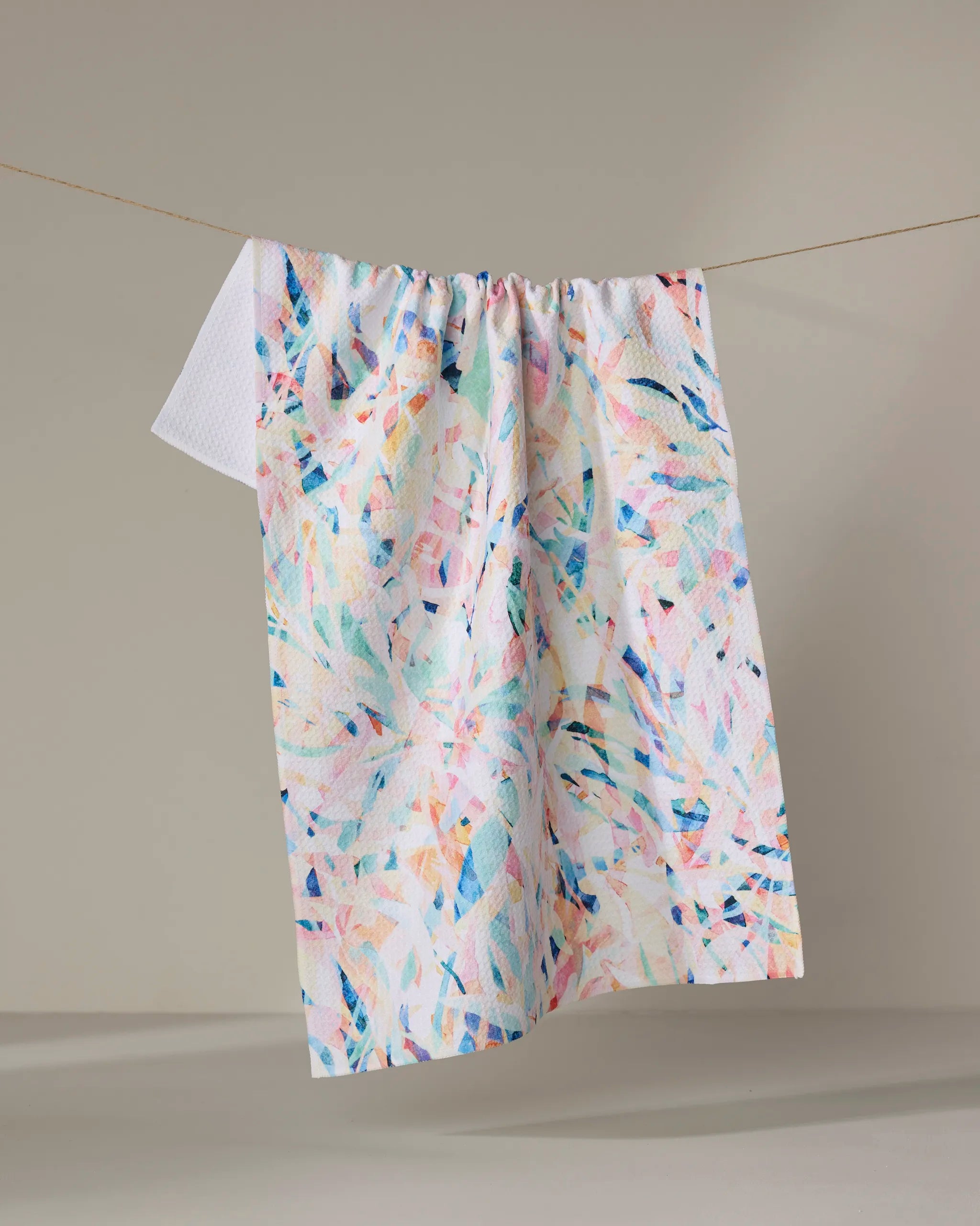 The "Abstract Jungle" tea towel by Rachel Parker, featuring pastel pinks, blues, yellows, and whites in brushstroke designs, hangs from a thin clothesline against a plain neutral background.