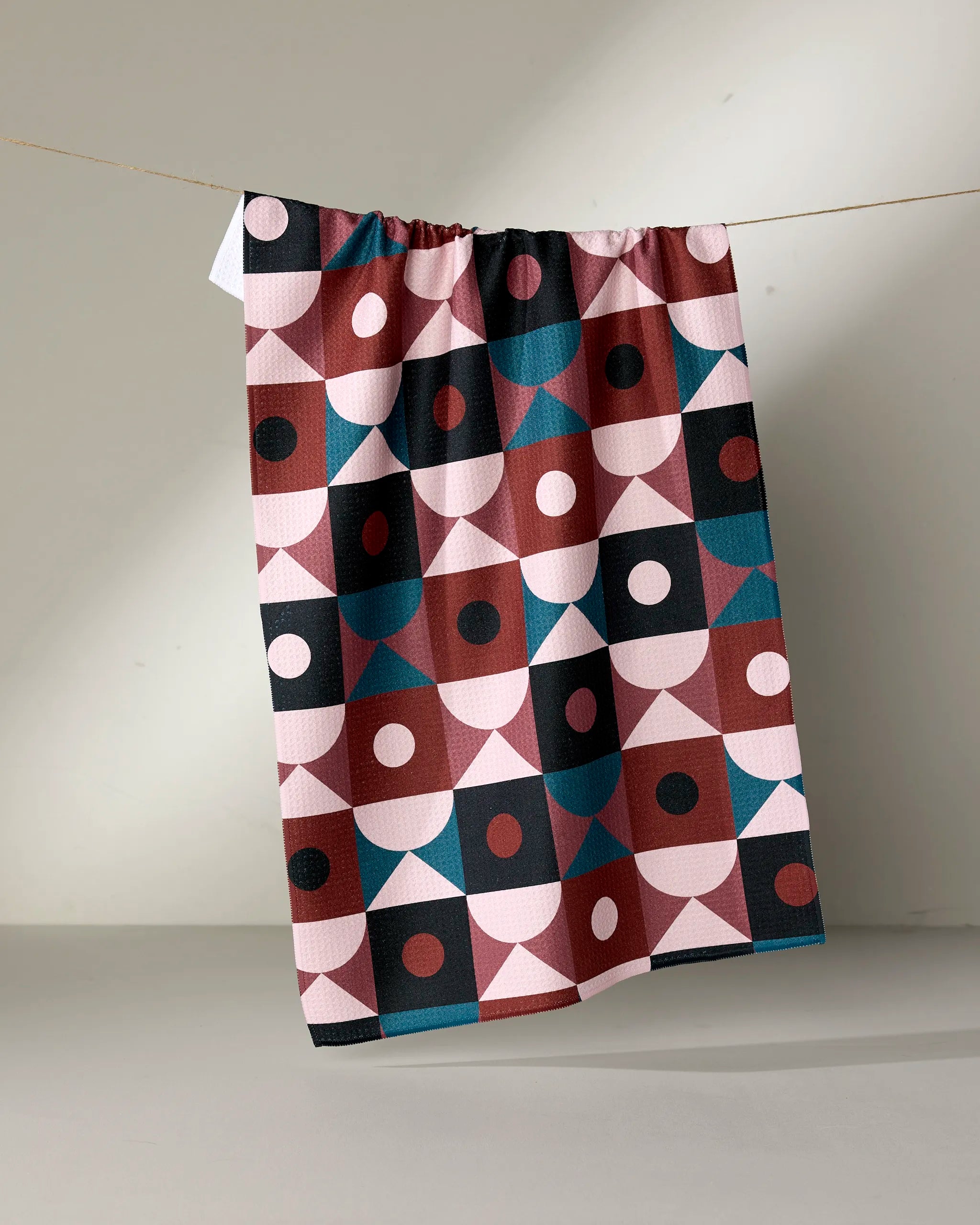 The "Abstract Retro" tea towel by Jennifer Bouron, featuring a geometric pattern in pink, burgundy, blue, and black, hangs on a string against a neutral background.