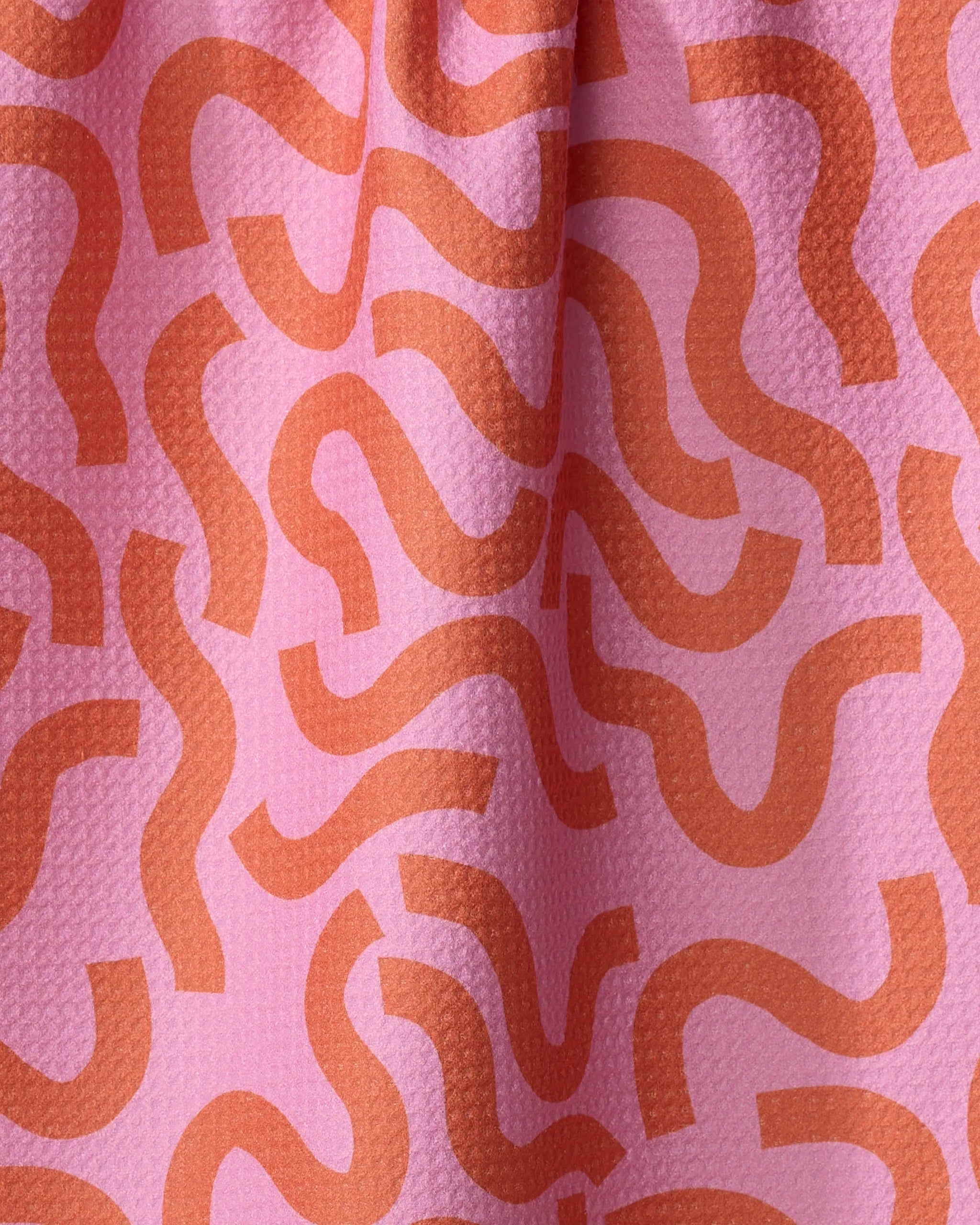 A close-up of the Jennifer Bouron "Abstract Snake" kitchen towel shows pink recycled fabric with an orange, wavy pattern, a slightly bumpy texture, and gentle folds.