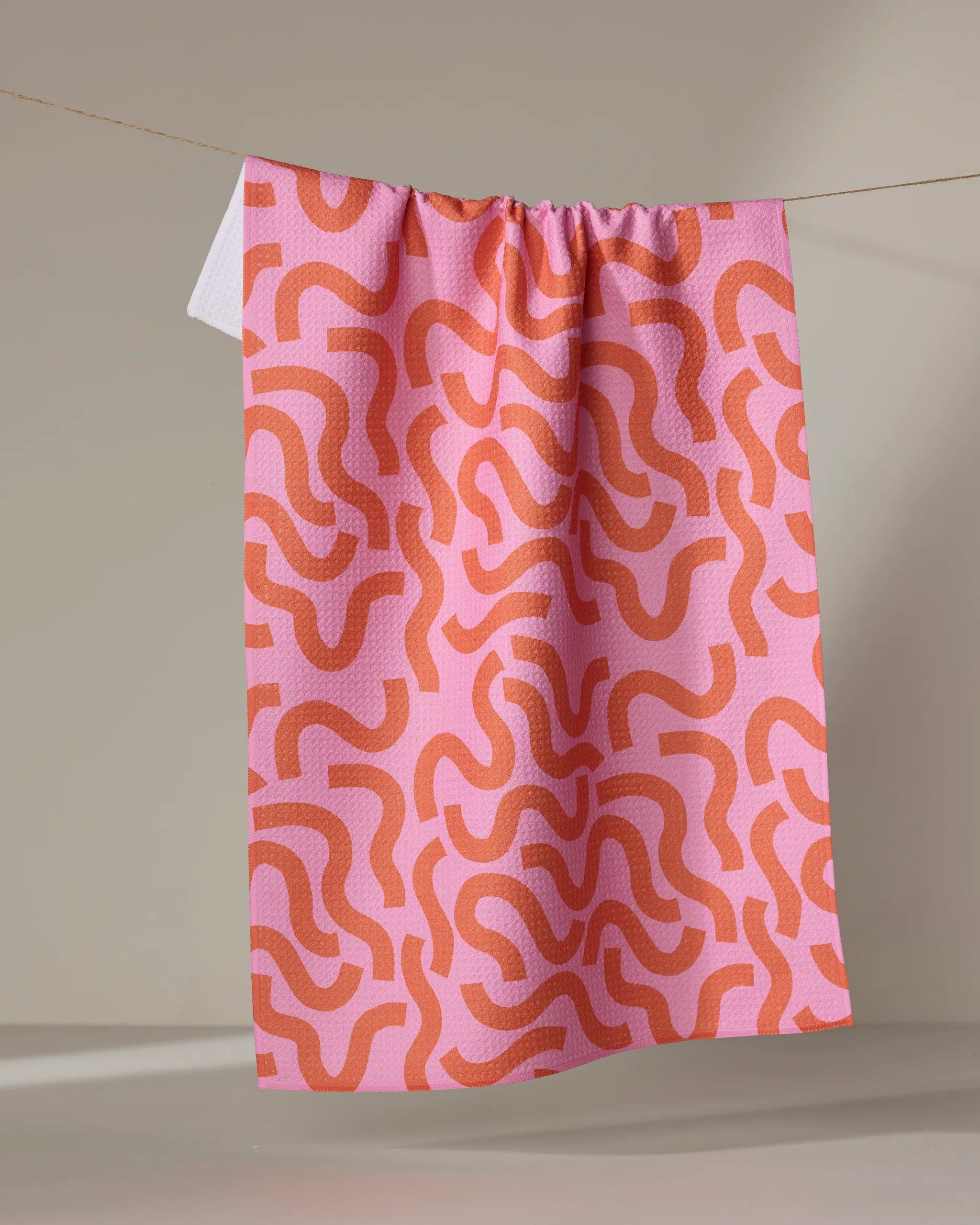 The Abstract Snake kitchen towel by Jennifer Bouron, featuring a pink and orange wavy design, hangs on a string against a neutral background.