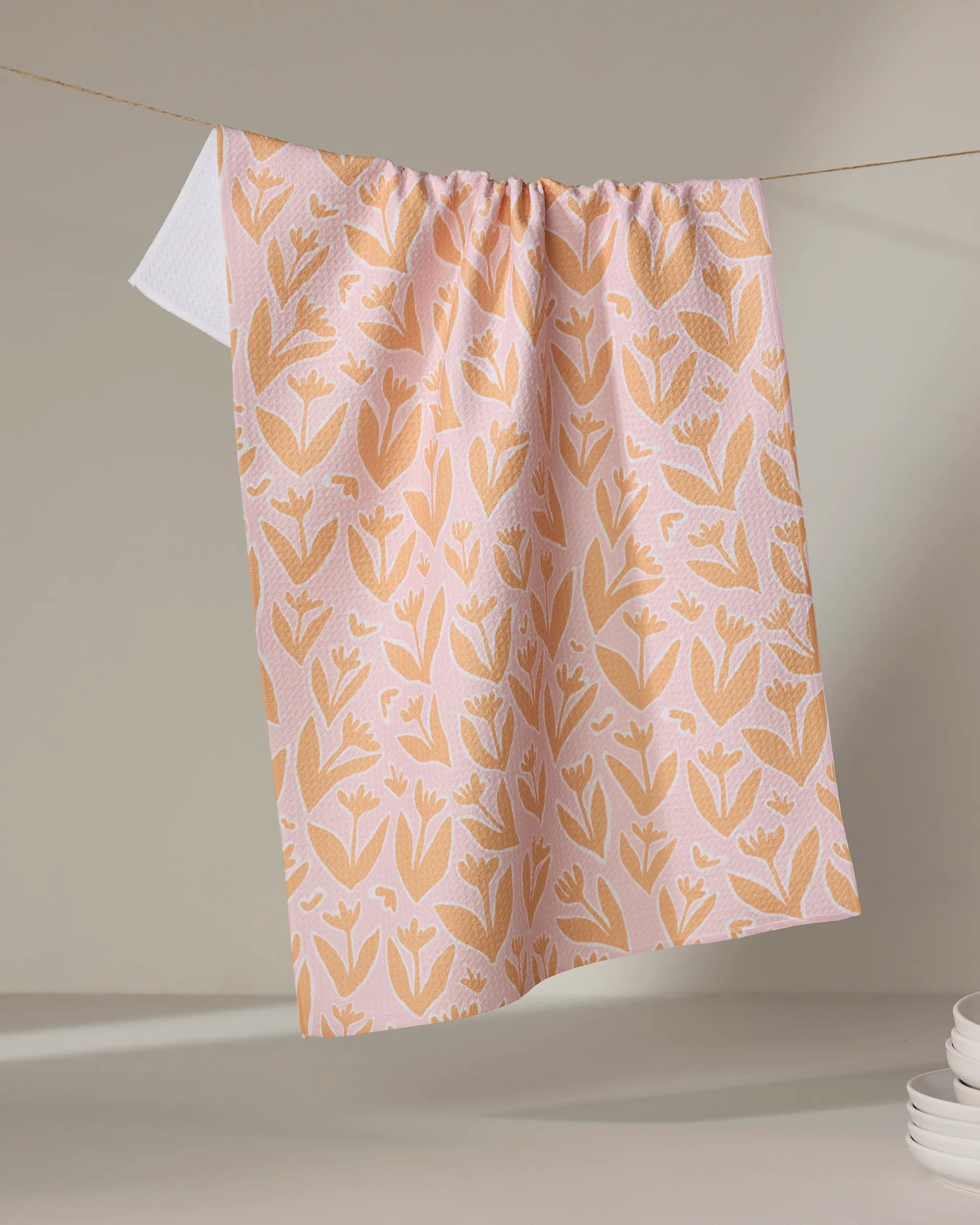 The Kayla Ann "Abstract Tulips" tea towel, featuring a pink and white floral pattern, hangs on a string against a neutral background with stacked white dishes in the lower right corner.