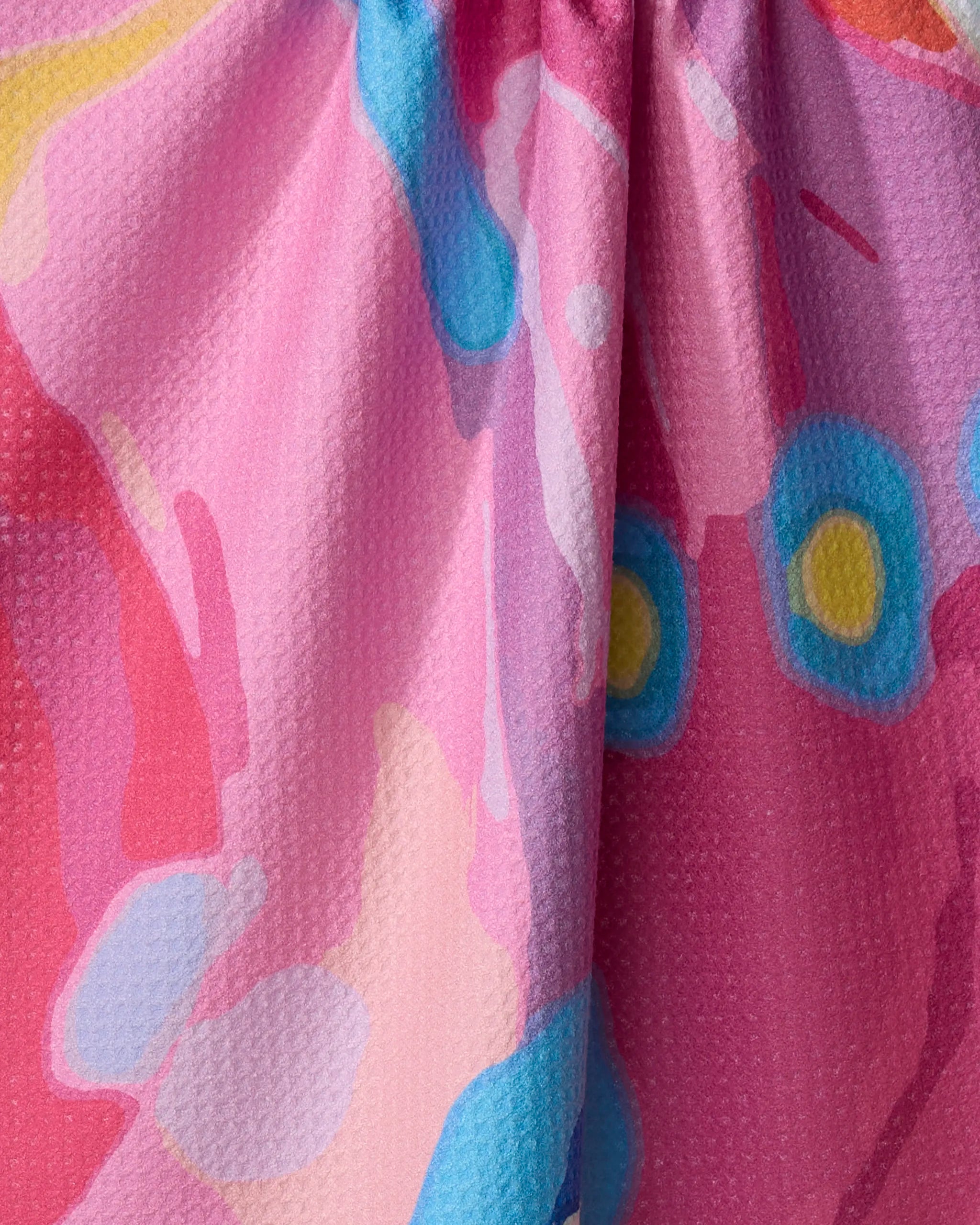 Close-up of Sareka Unique's Abundance tea towel in pink, made from recycled materials. Features abstract blue, yellow, and red patterns with subtle folds near the top that create gentle shadows.