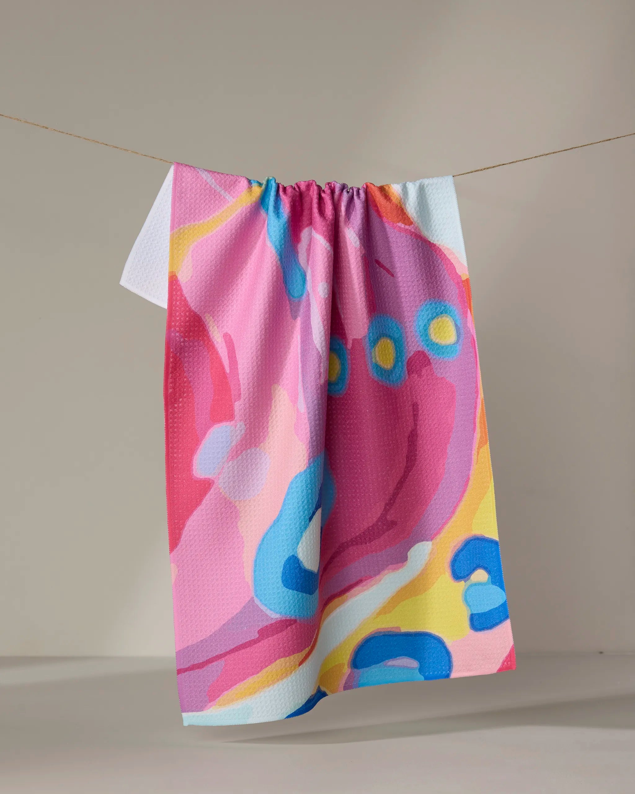 The "Abundance" towel by Sareka Unique, featuring quick-drying fabric and vibrant abstract patterns in pink, blue, yellow, and white, hangs on a string against a neutral beige background.