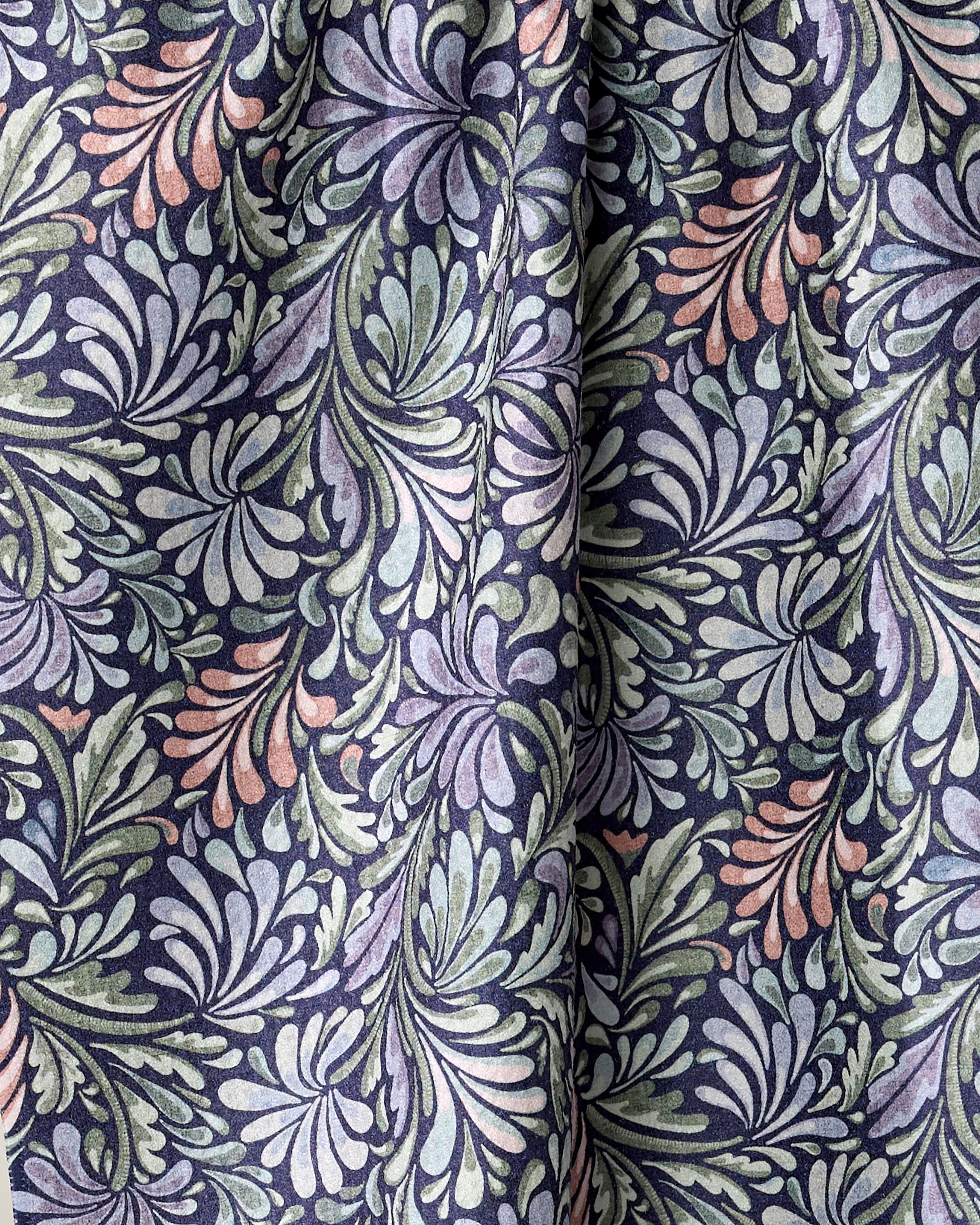 A close-up of the Kris Ruff Acanthus kitchen tea towel, crafted from recycled materials and showcasing swirling leafy patterns in muted green, purple, and peach on a dark background. The fabric is softly gathered with visible folds.