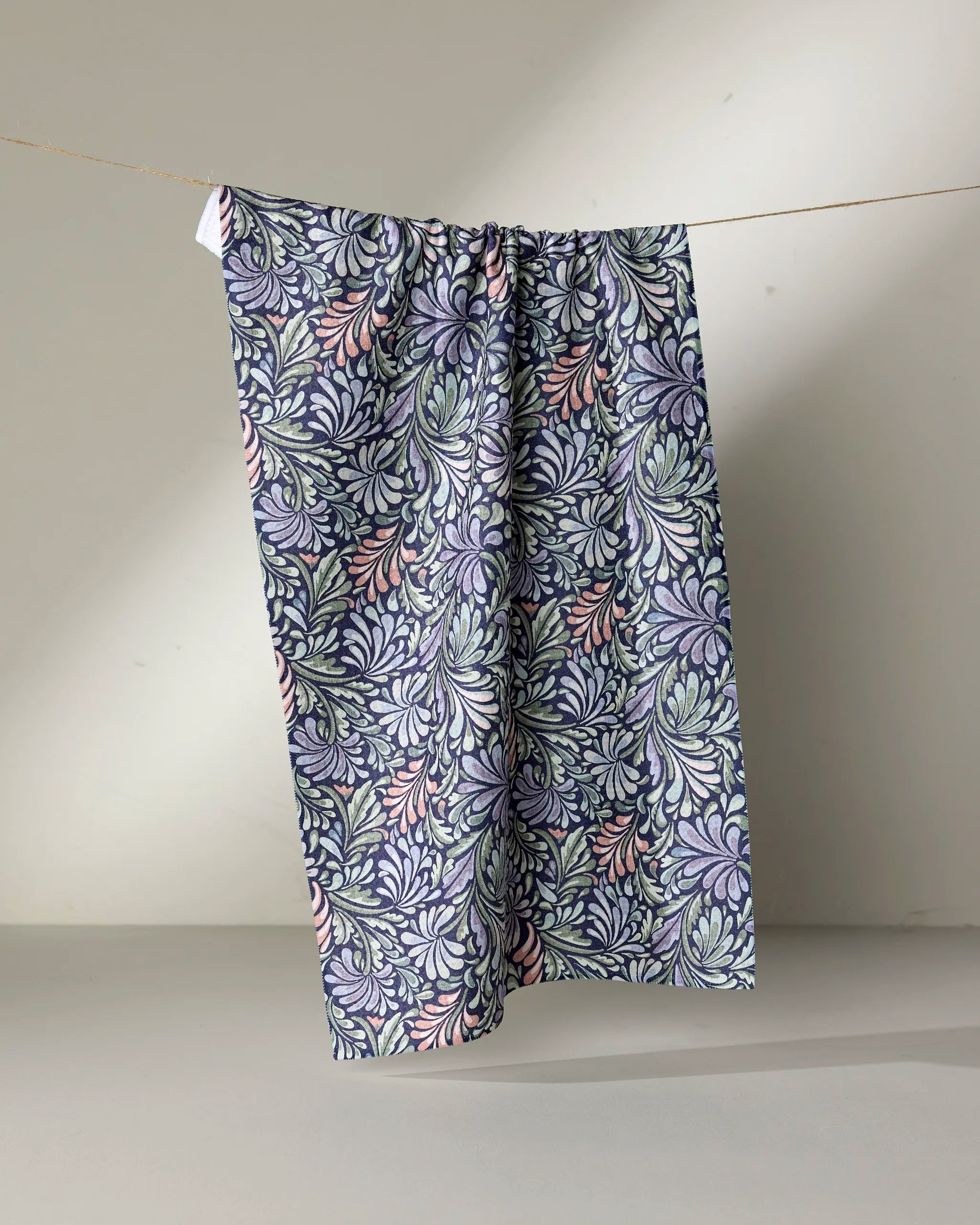 Acanthus by Kris Ruff is a patterned fabric made from recycled materials, featuring a blue, green, and pink floral design, shown hanging on a clothesline against a light background.