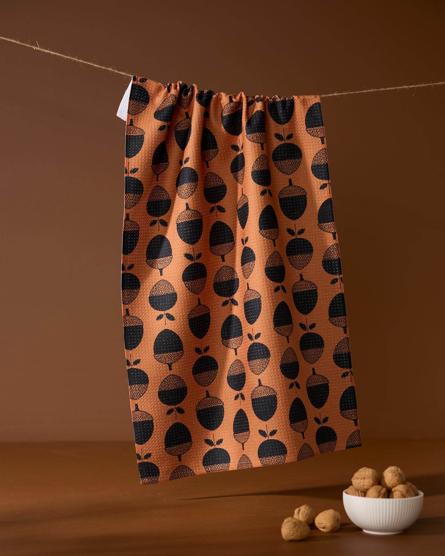 The Acorn Noir kitchen tea towel by Geometry, featuring a black acorn pattern and made from recycled materials, hangs on a string above a small white bowl of walnuts and loose nuts, set against a brown background.
