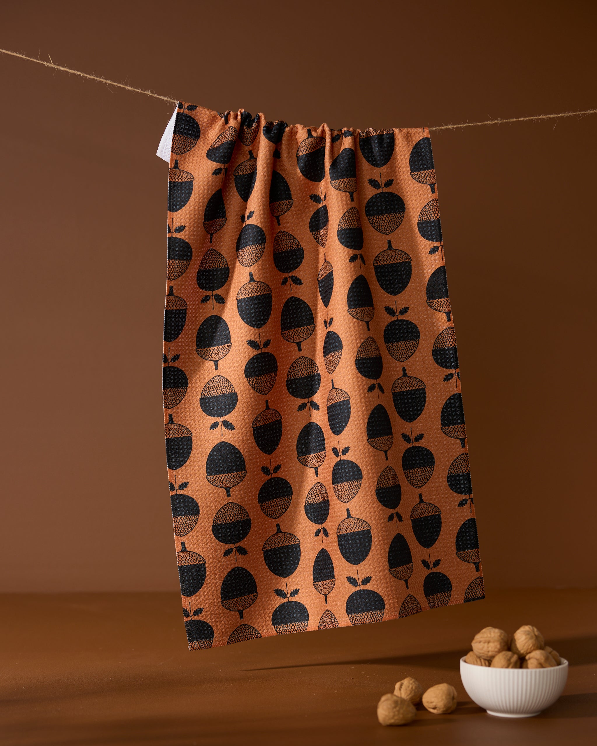 The Acorn Noir kitchen tea towel by Geometry, featuring a black acorn pattern and made from recycled materials, hangs on a string above a small white bowl of walnuts and loose nuts, set against a brown background.