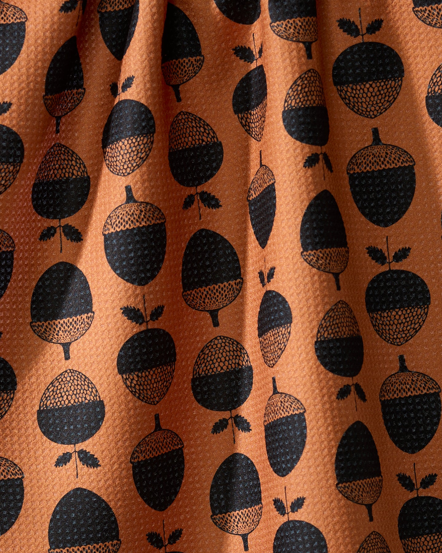 The Geometry Acorn Noir kitchen tea towel features a textured orange fabric with evenly spaced rows of black acorns and leaves. Gentle folds add shadows and highlights to its ultra-absorbent design.