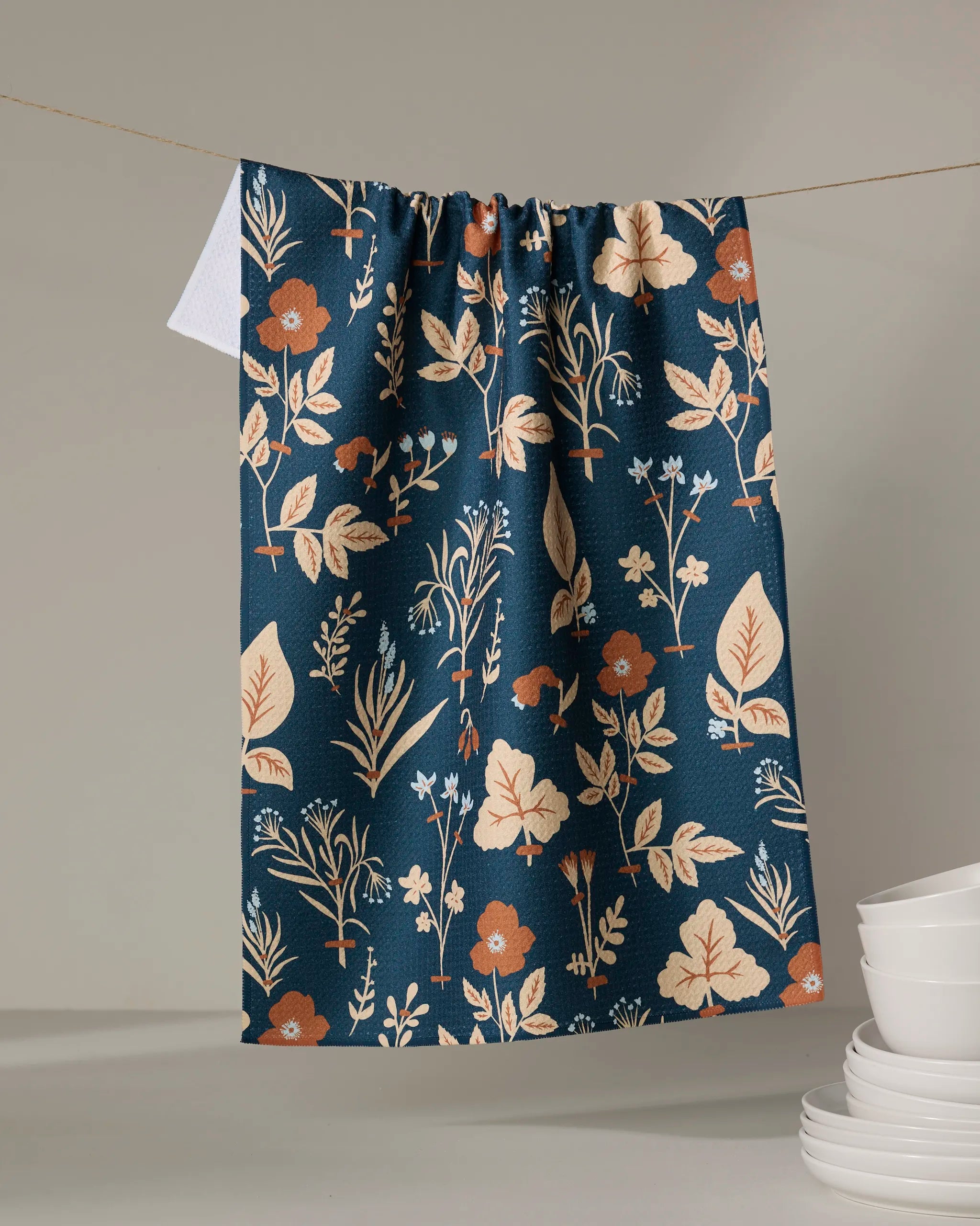 The Aemilius tea towel by Danelys Sidron, featuring a dark blue background with beige and rust floral accents, hangs on a line. A white cloth is partially visible behind it, with stacked white dishes nearby on a gray surface.