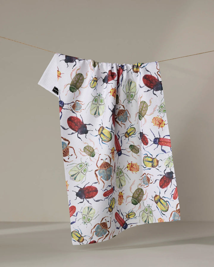 The "African Beetles" recycled kitchen towel by Taryn Brennan, featuring colorful beetle illustrations, hangs on a clothesline against a neutral backdrop.