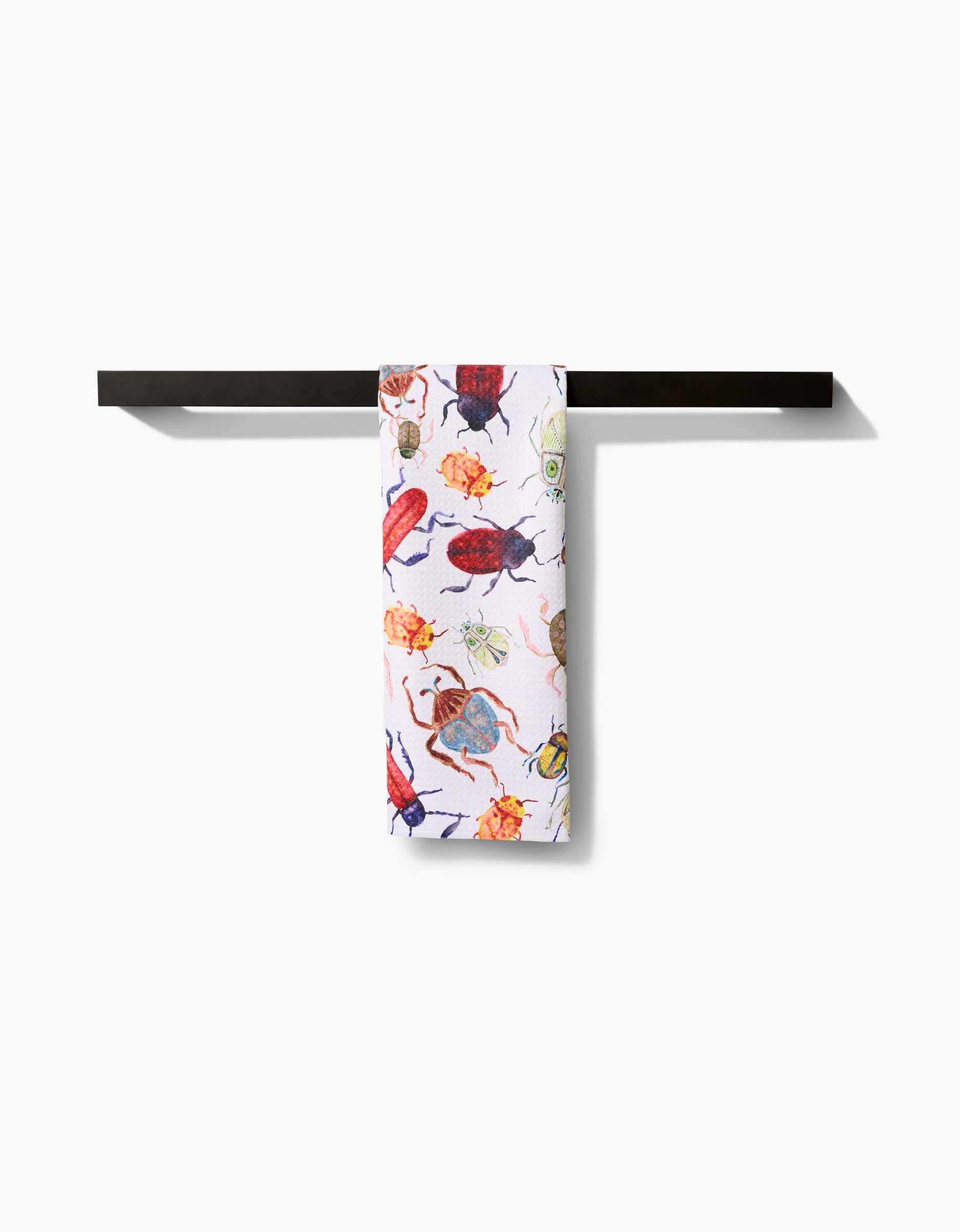 The Taryn Brennan African Beetles tea towel, featuring colorful illustrated beetles and insects on white, is neatly draped over a sleek black towel bar against a white background.