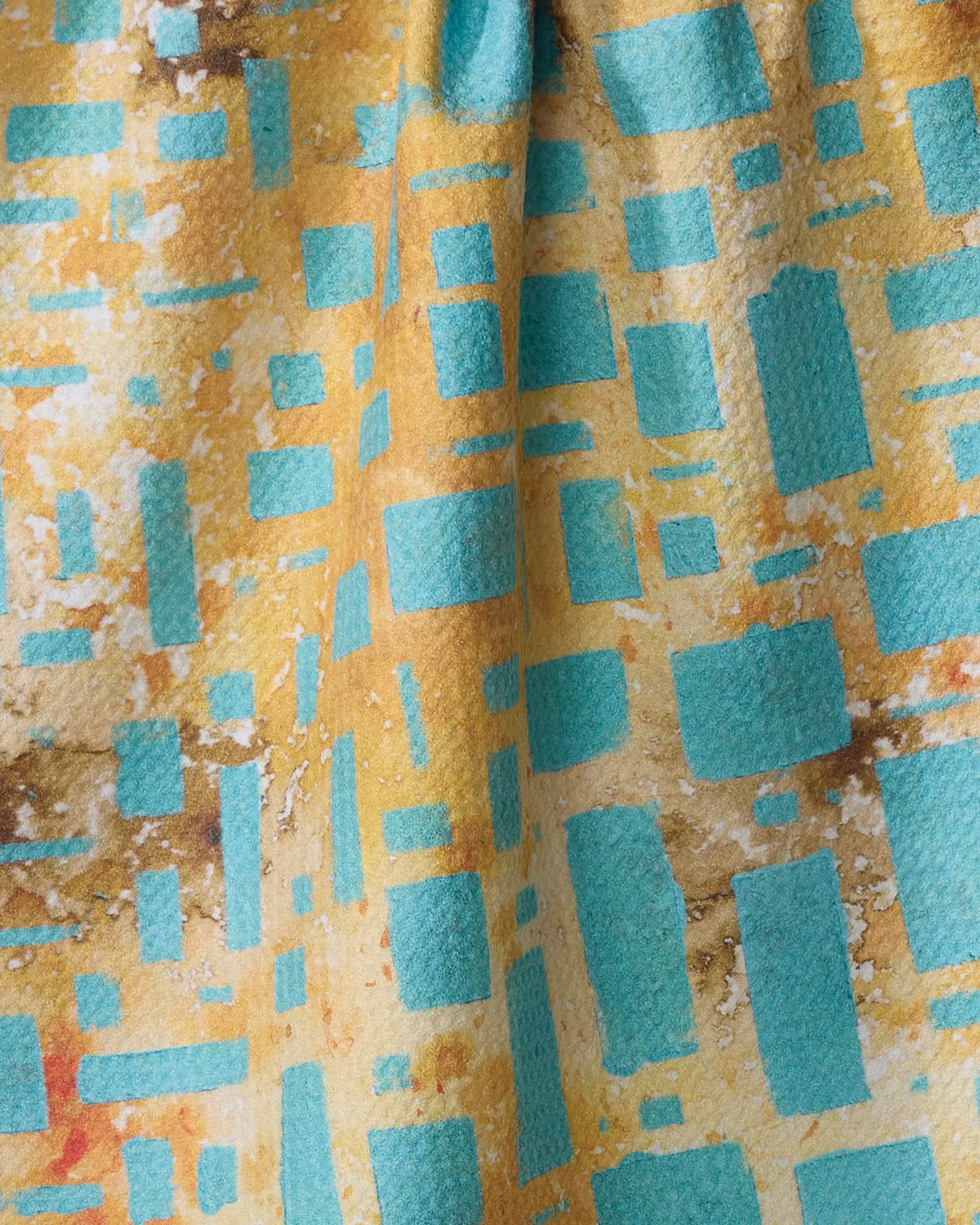 A close-up of Taryn Brennan's African Blue Geometry fabric, showcasing recycled material with turquoise rectangles and squares in a geometric pattern on a yellow and brown background, highlighted by areas of light and shadow.