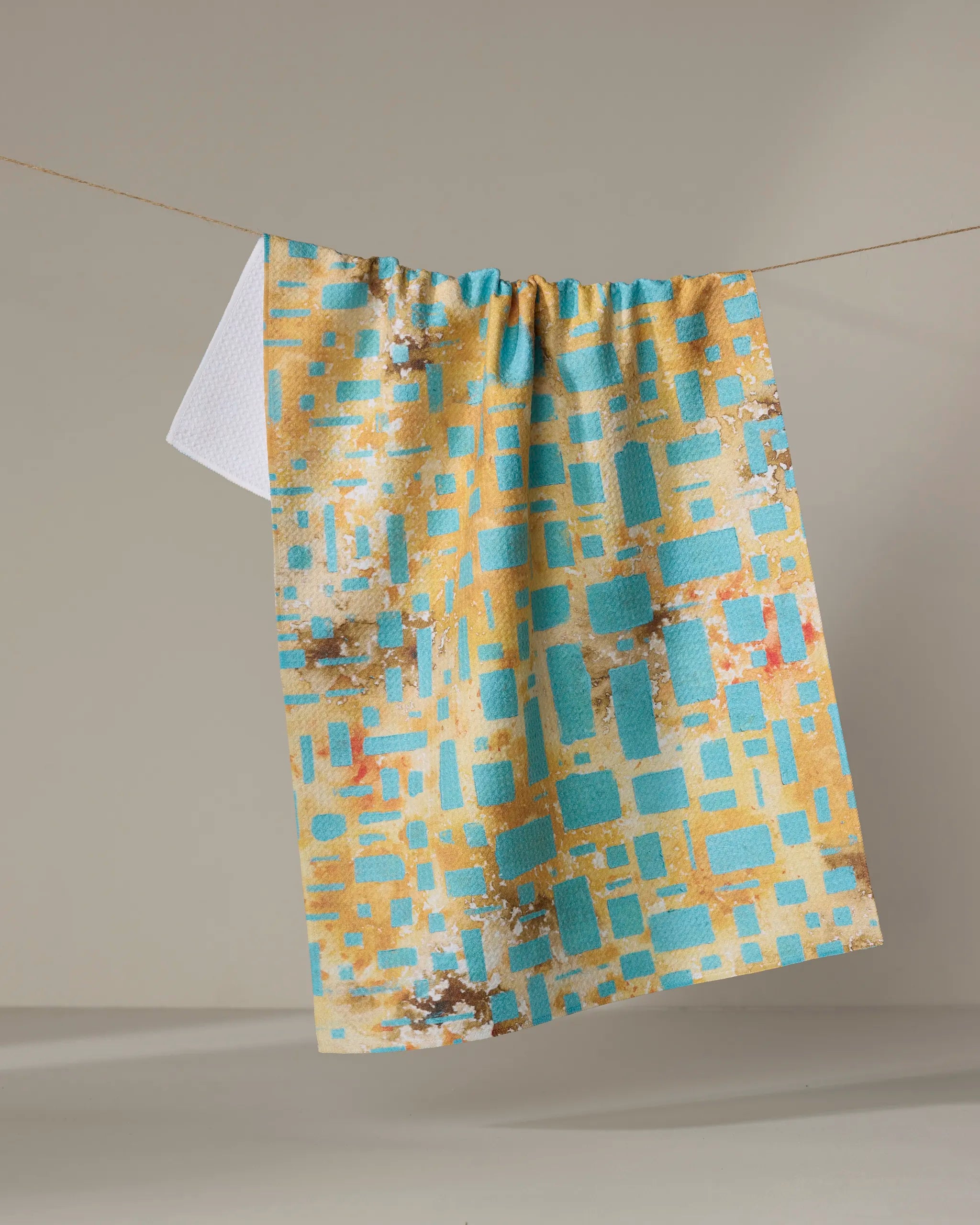 The "African Blue Geometry" tea towel by Taryn Brennan, made from recycled materials, showcases turquoise, gold, and beige geometric patterns with a small white corner detail. It’s pictured hanging against a neutral background.
