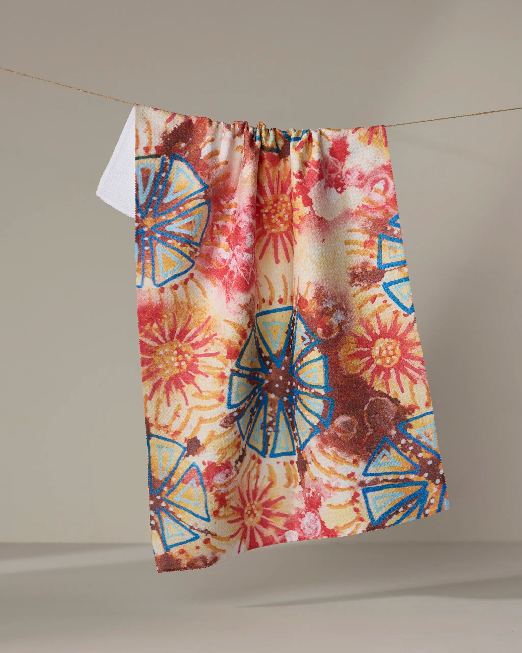The "African Loose Circles" towel by Taryn Brennan, featuring vibrant blue geometric and orange-yellow floral patterns, hangs on a clothesline against a neutral background.