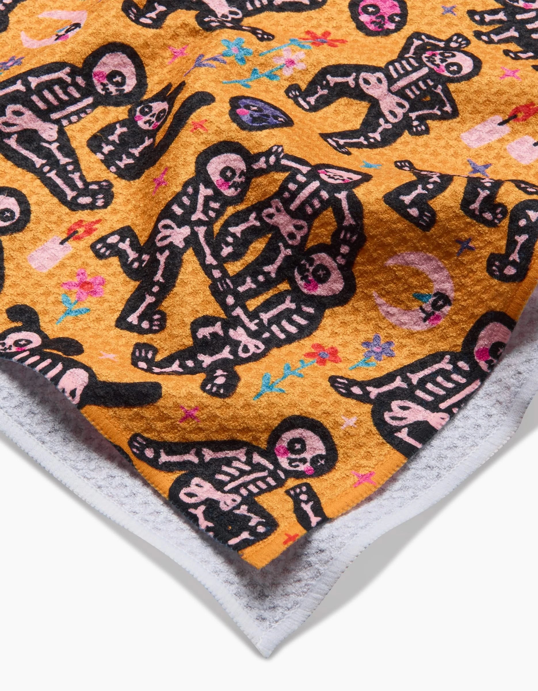 A close-up of the So Lazo After Party tea towel, featuring black and pink cartoon skeletons with colorful flowers on yellow-orange, soft white edges, and made from ultra-absorbent recycled materials.