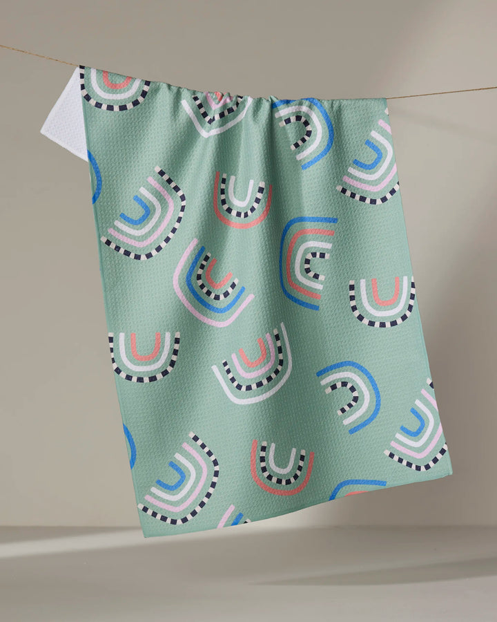 The "After the Rain" tea towel by Claire Iglesias features a light green textured fabric with colorful pink, white, blue, and black semi-circles and arches, hanging on a line against a neutral background.