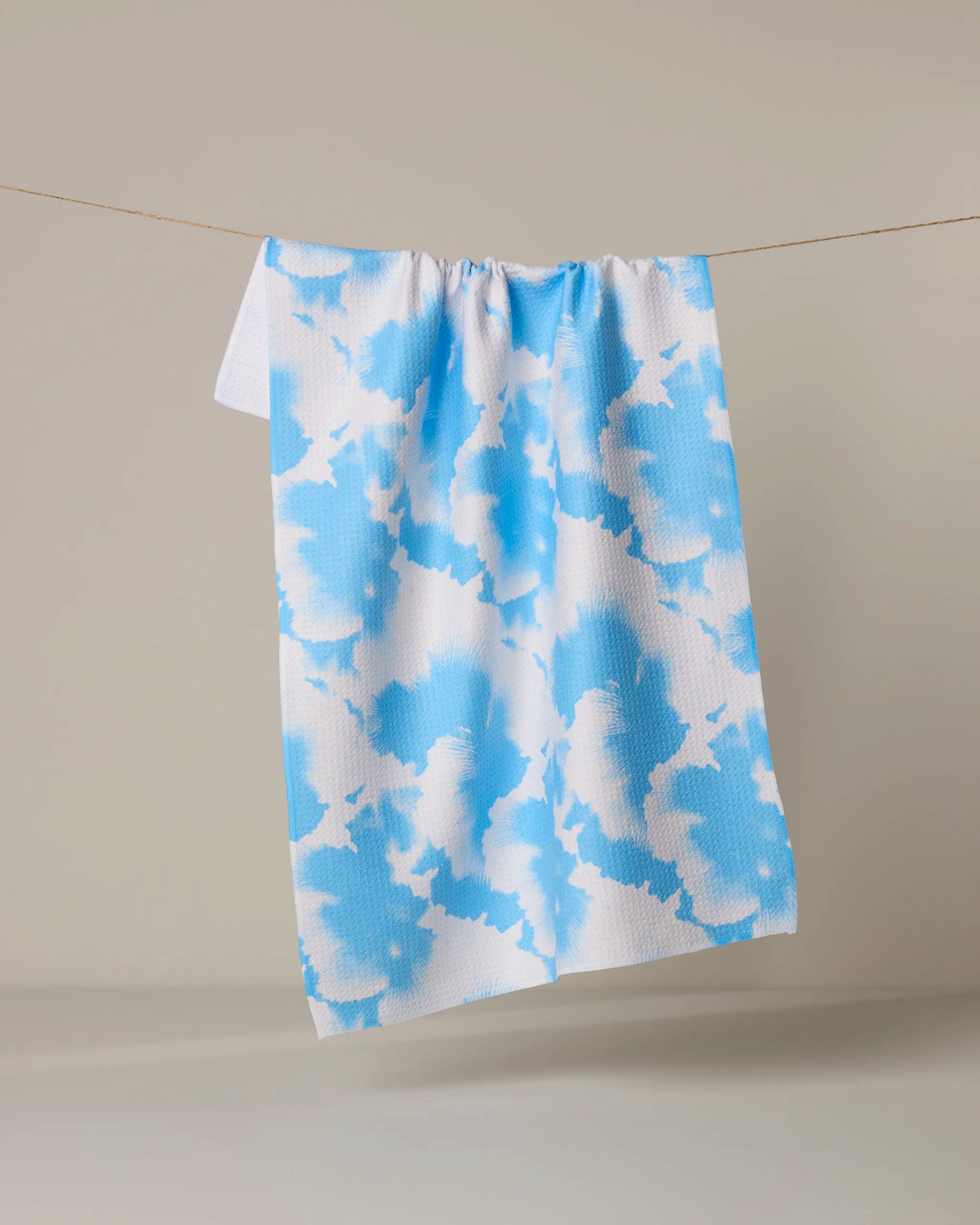 The Alicia towel by Geometry, featuring a quick-drying abstract blue-on-white cloud design, hangs on a thin string against a plain beige background.
