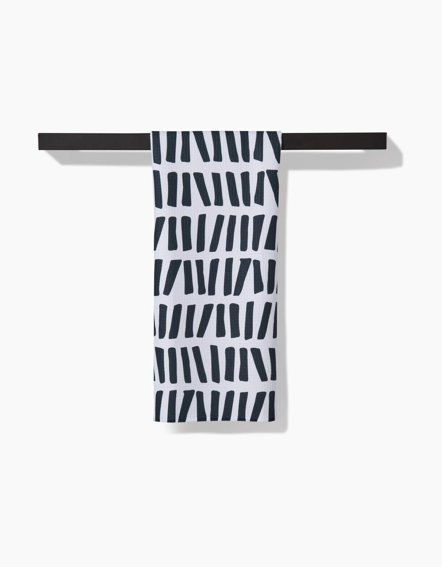 The GEOMETRY All Lined Up white ultra-absorbent tea towel, featuring irregular black lines, hangs neatly over a slim black towel bar against a plain white background.