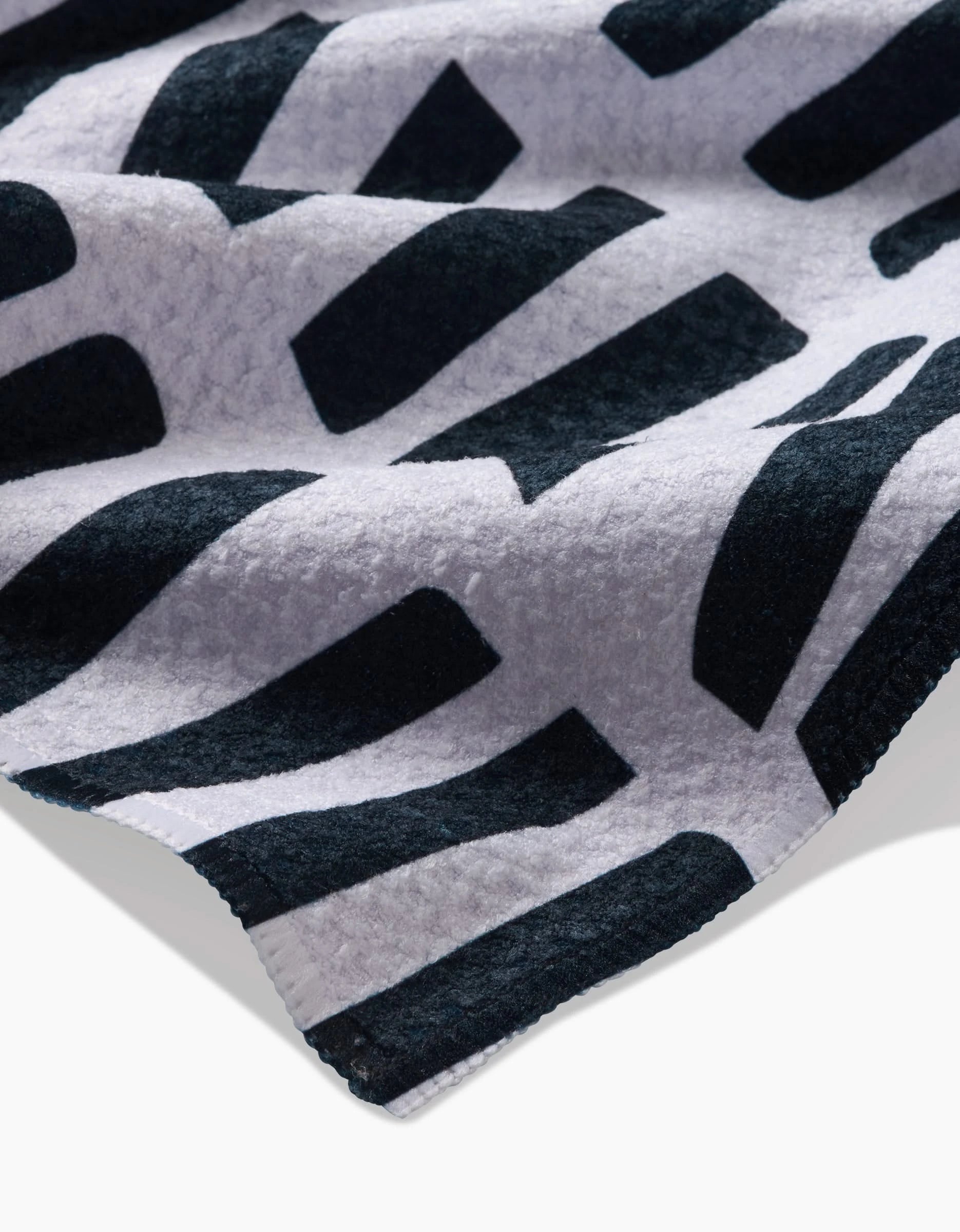 A close-up of GEOMETRYs All Lined Up shows its soft, thick fabric made from recycled materials with striking black and white geometric patterns. A curled edge reveals the eco-friendly materials texture.