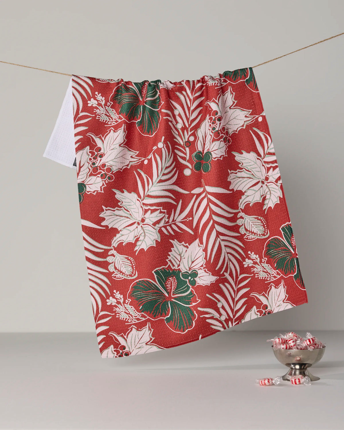 Aloha Winters by Geometry is an absorbent dish towel featuring a red and white floral design with green accents, shown hanging on a line. Below, a silver bowl holds unwrapped peppermint candies, with several candies scattered nearby—adding both charm and function to any kitchen scene.