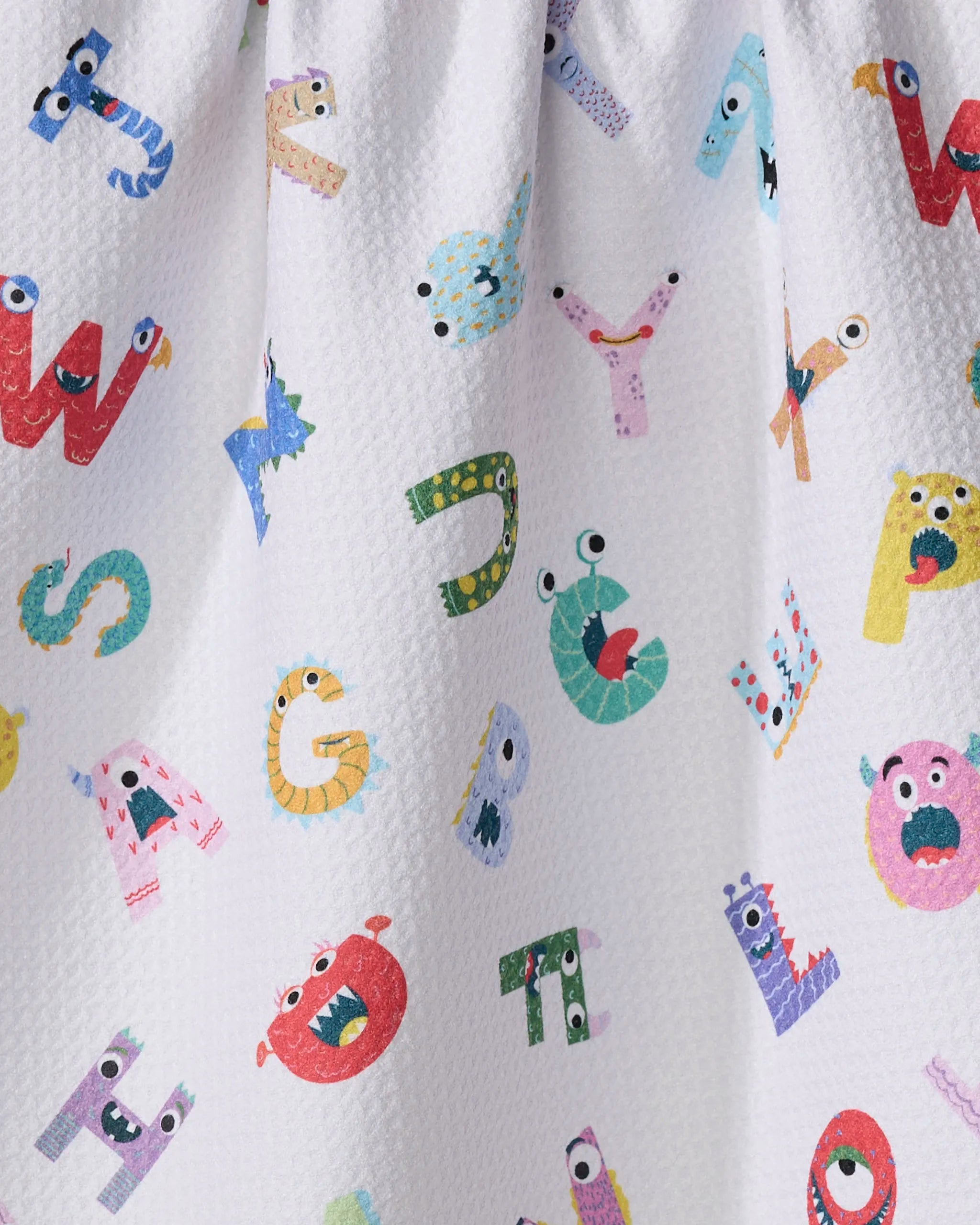 The "Alphabet Monsters" tea towel by Vaishali Design Studio features colorful cartoon letters with playful monster faces, each with unique eyes and mouths, printed on a white, textured, ultra-absorbent fabric for a whimsical look.