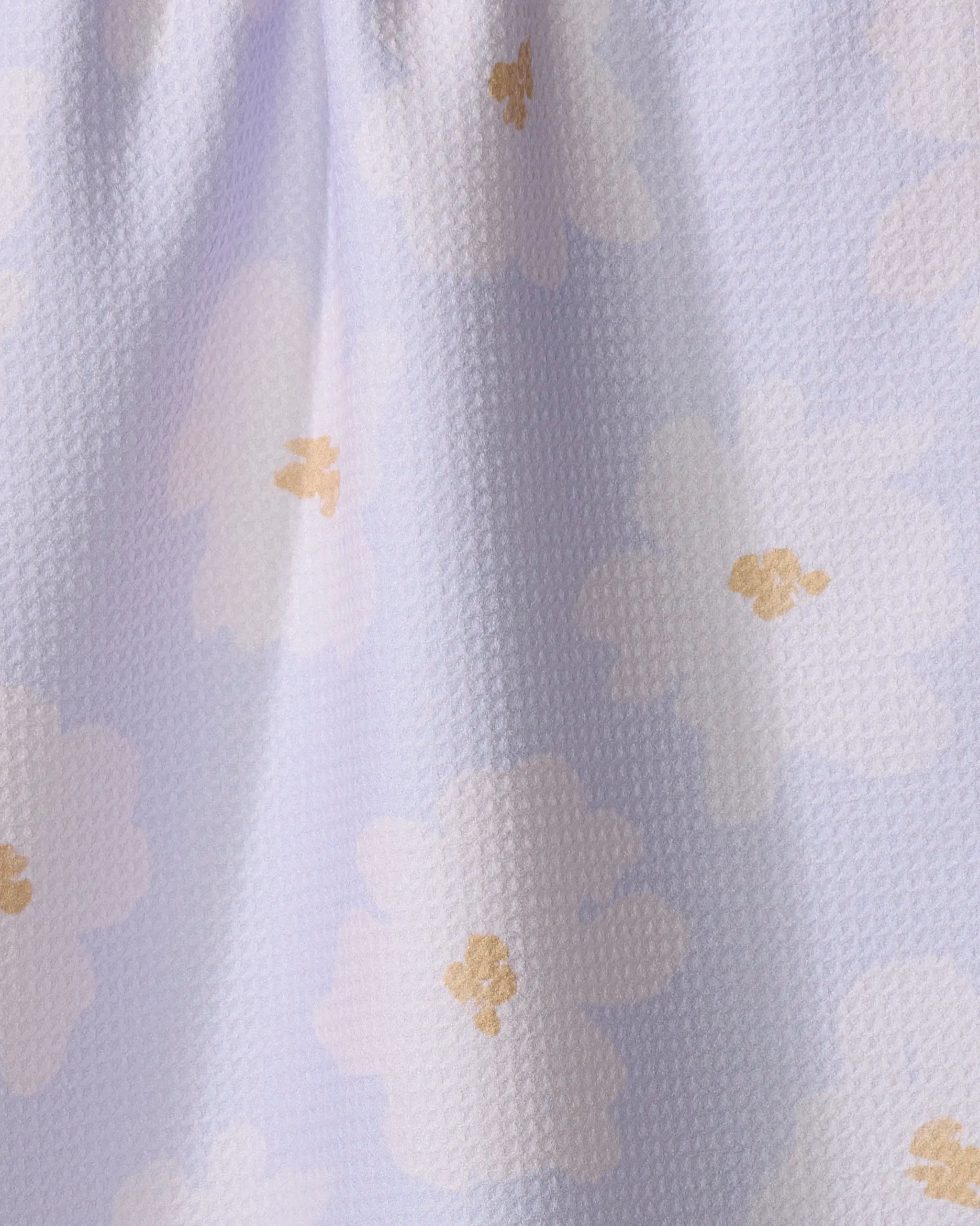 A close-up of Alpine Blooms by Mandy Arroyo shows textured white recycled fabric, featuring pale lavender flowers with yellow centers. The softly draped kitchen towel forms gentle folds and shadows.