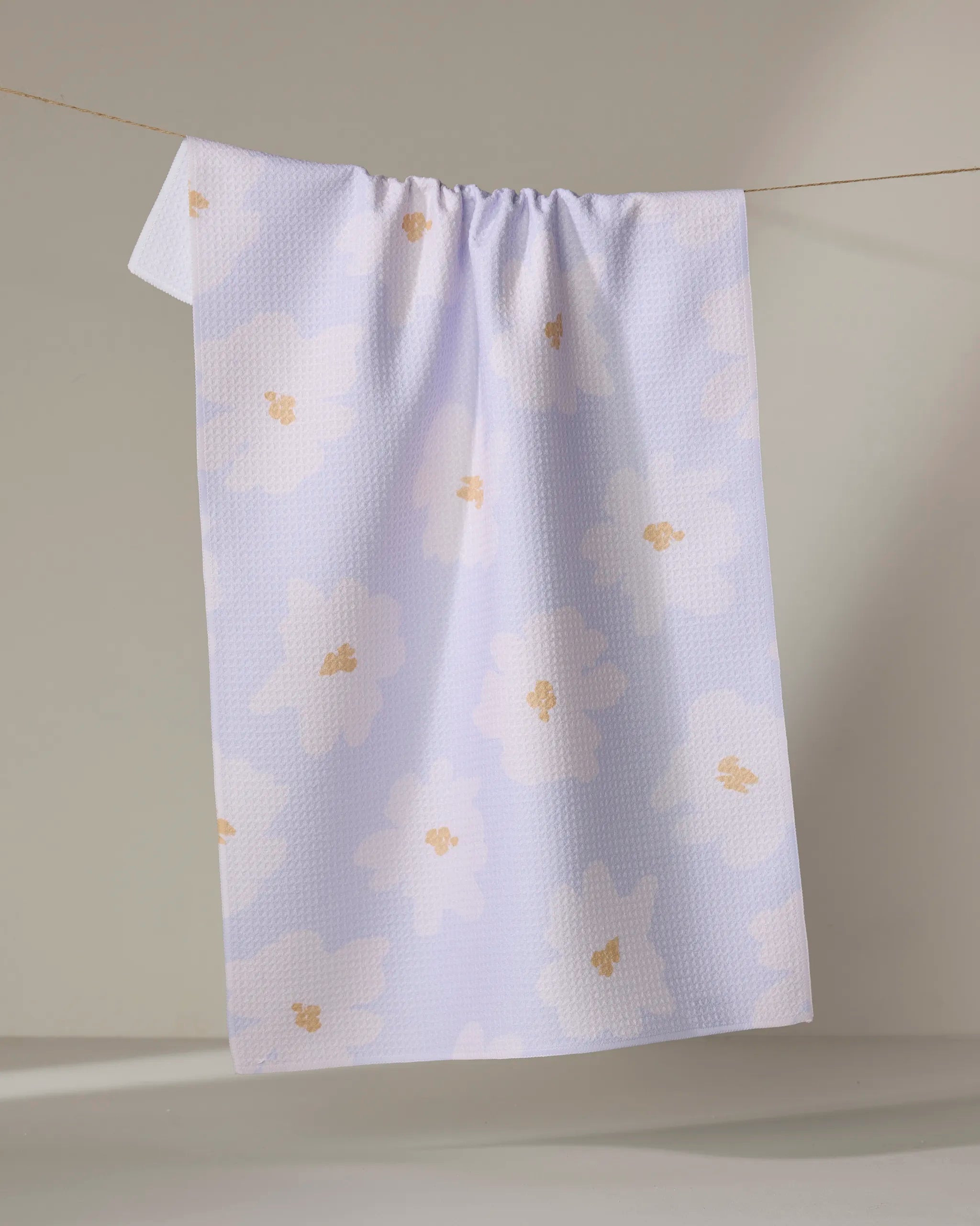 The Mandy Arroyo "Alpine Blooms" tea towel, featuring a textured pattern and bold yellow and white floral designs on light purple, hangs neatly on a thin string against a neutral background.