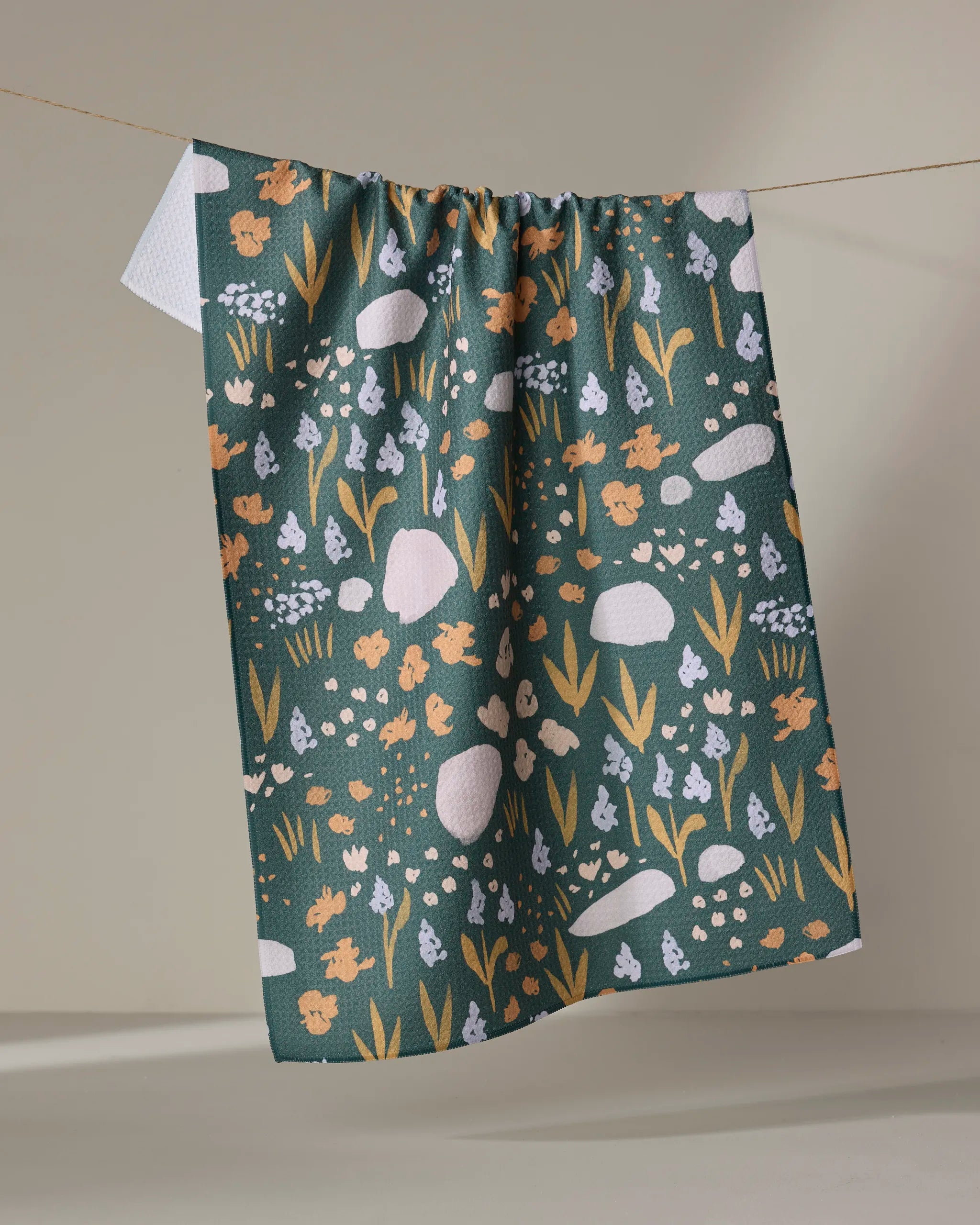 The Alpine Meadow Bold by Mandy Arroyo, a dark green ultra-absorbent kitchen tea towel featuring whimsical white, yellow, and light blue floral and stone patterns, hangs on a clothesline against a neutral background.