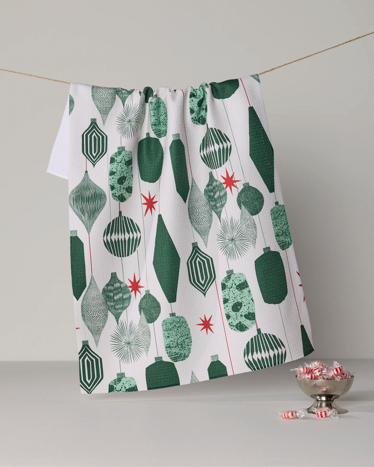 The Alpine Revelry tea towel from Geometry, featuring green and red geometric ornament patterns on crisp white, hangs elegantly on a string. Made from quick-drying, recycled materials, it complements the festive display beneath—a silver bowl brimming with peppermint candies, some playfully scattered across the snowy-white surface.