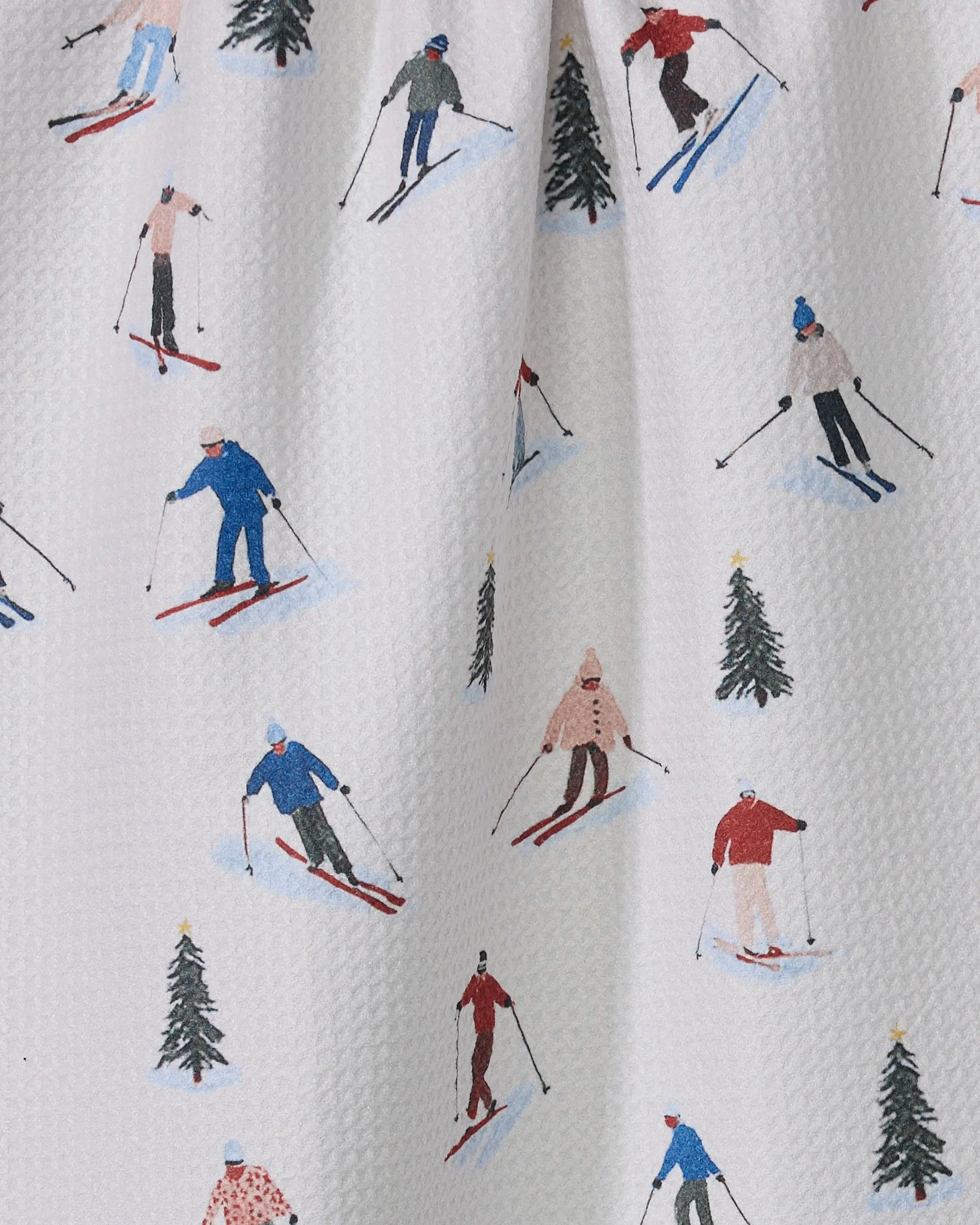 The Geometry "Alpine Run" tea towel features skiers in colorful winter outfits among green pine trees on a textured white background. Made from recycled materials, it adds an eco-friendly touch to your kitchen.
