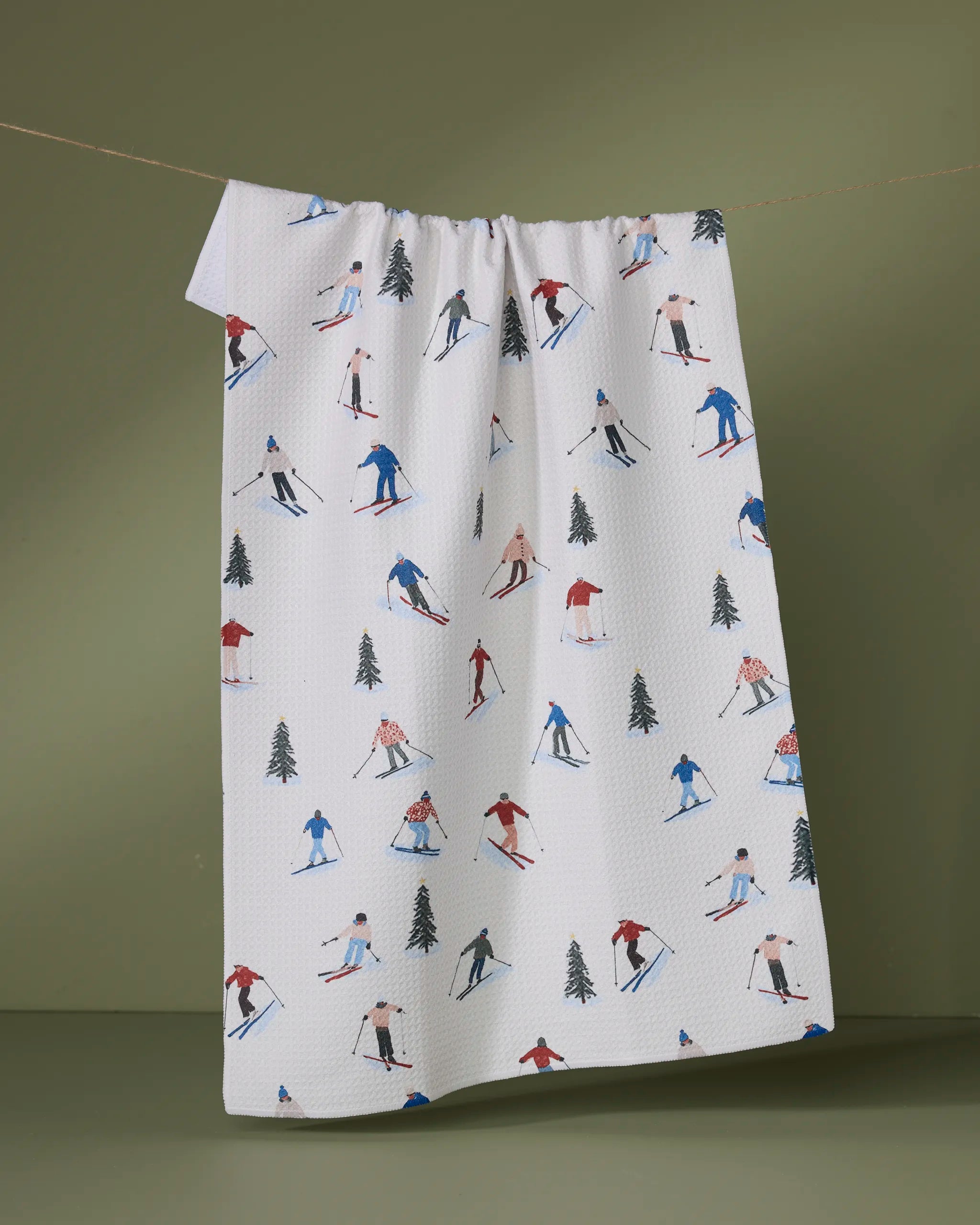 The "Alpine Run" kitchen towel by Geometry, crafted from recycled materials and featuring colorful skiers and green pine trees, hangs against a plain olive-green background.