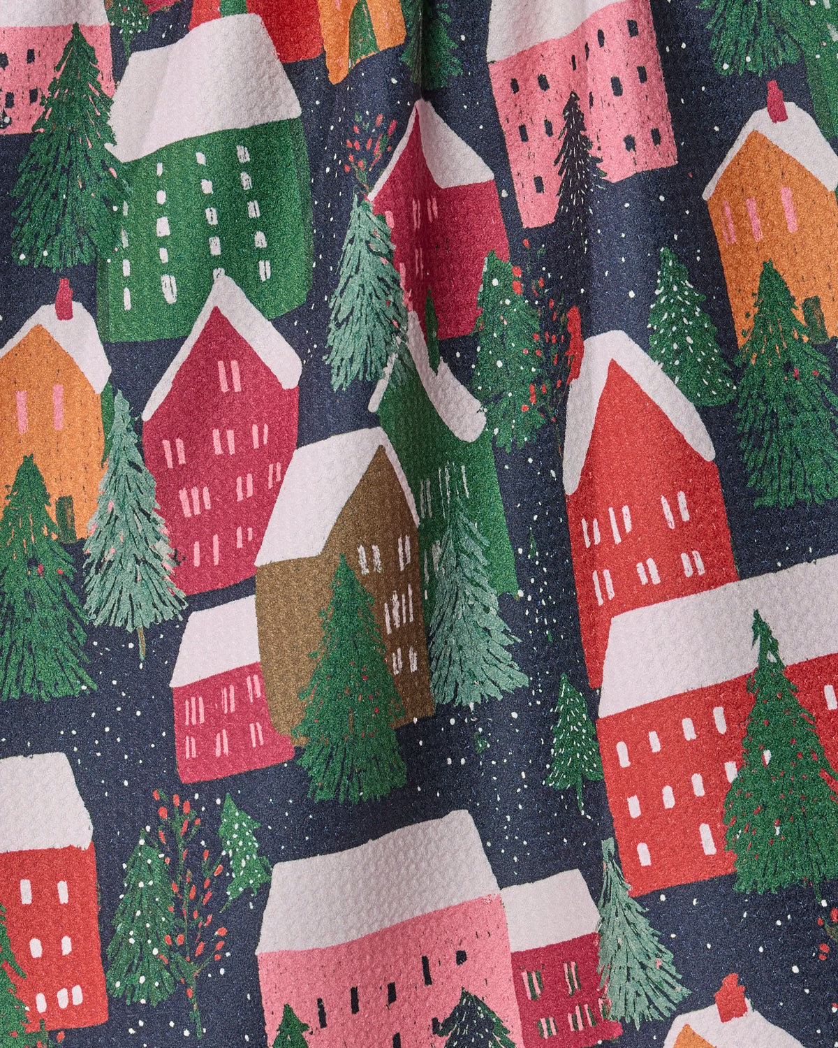 The “Alpine Village” tea towel by Geometry features a festive winter holiday village scene with colorful houses, green pine trees, and snow-covered roofs set against a dark blue, starry night—crafted from ultra-absorbent recycled materials.