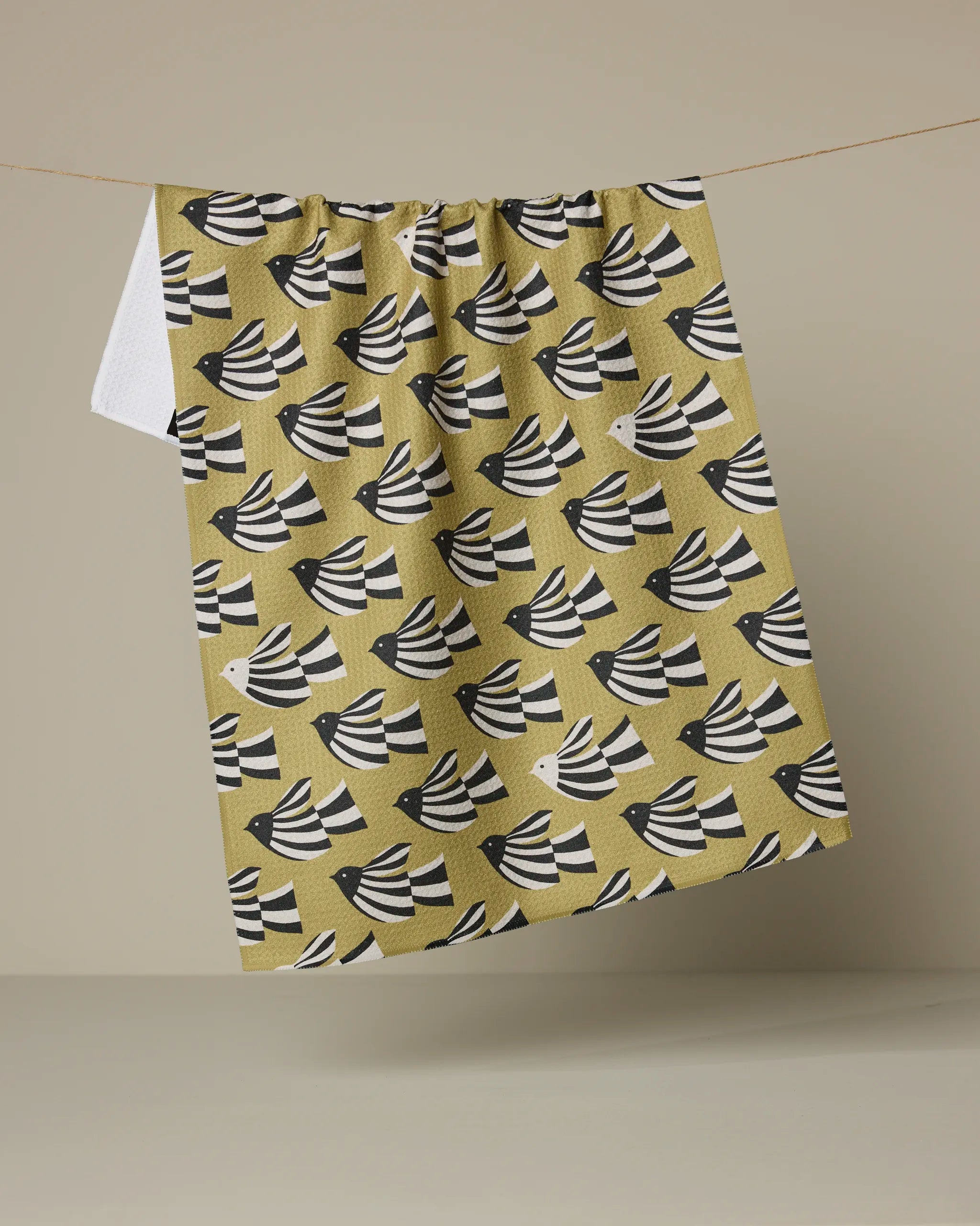 The Alva tea towel by Anna Christian, crafted from recycled materials, displays black and white bird motifs on a mustard yellow background and hangs on a clothesline against a beige backdrop.
