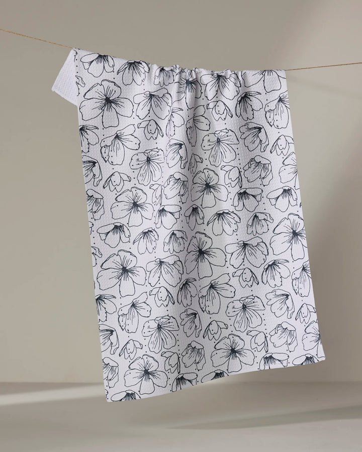 The "Always Blooming" recycled kitchen towel by Rosa Kichline, featuring a black floral line art pattern, hangs on a thin clothesline against a neutral beige background.