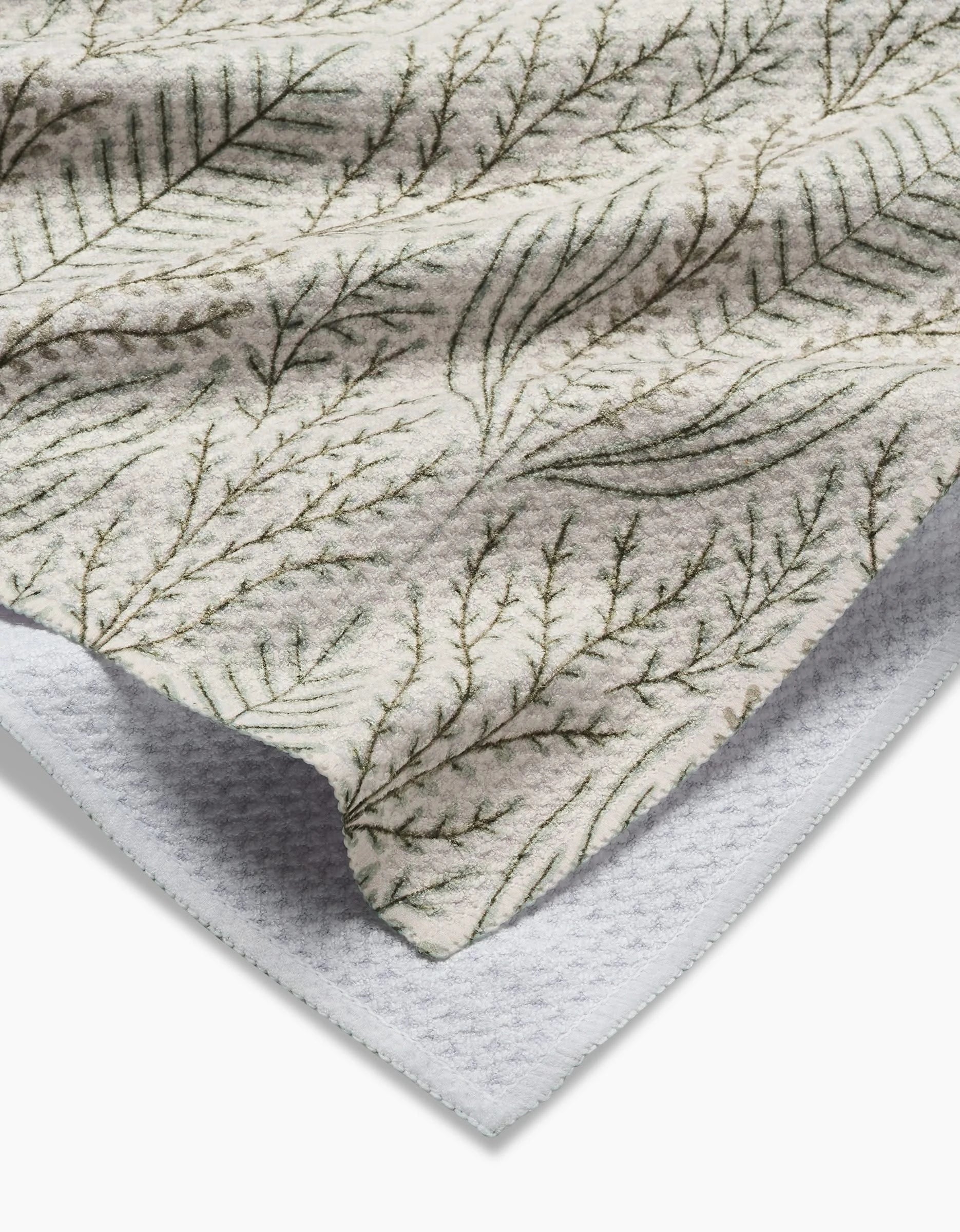 A close-up of the Amberlin Tea Towel by Amy MacCready, crafted from Geoweave fabric, displays a leafy green pattern on beige, layered over a white textured surface.