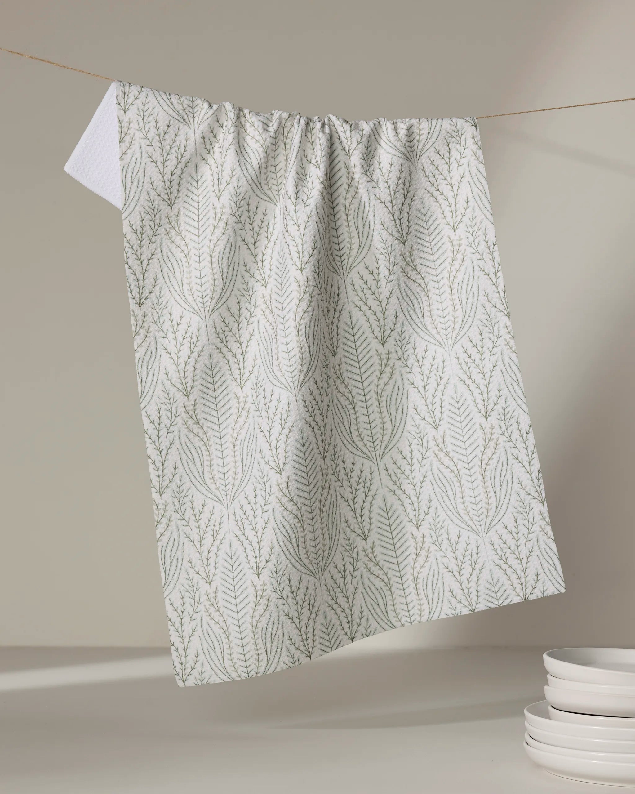 The Amberlin tea towel by Amy MacCready, made from recycled material with a subtle green leafy pattern, hangs on a string against a beige background, next to a stack of white plates on the right.