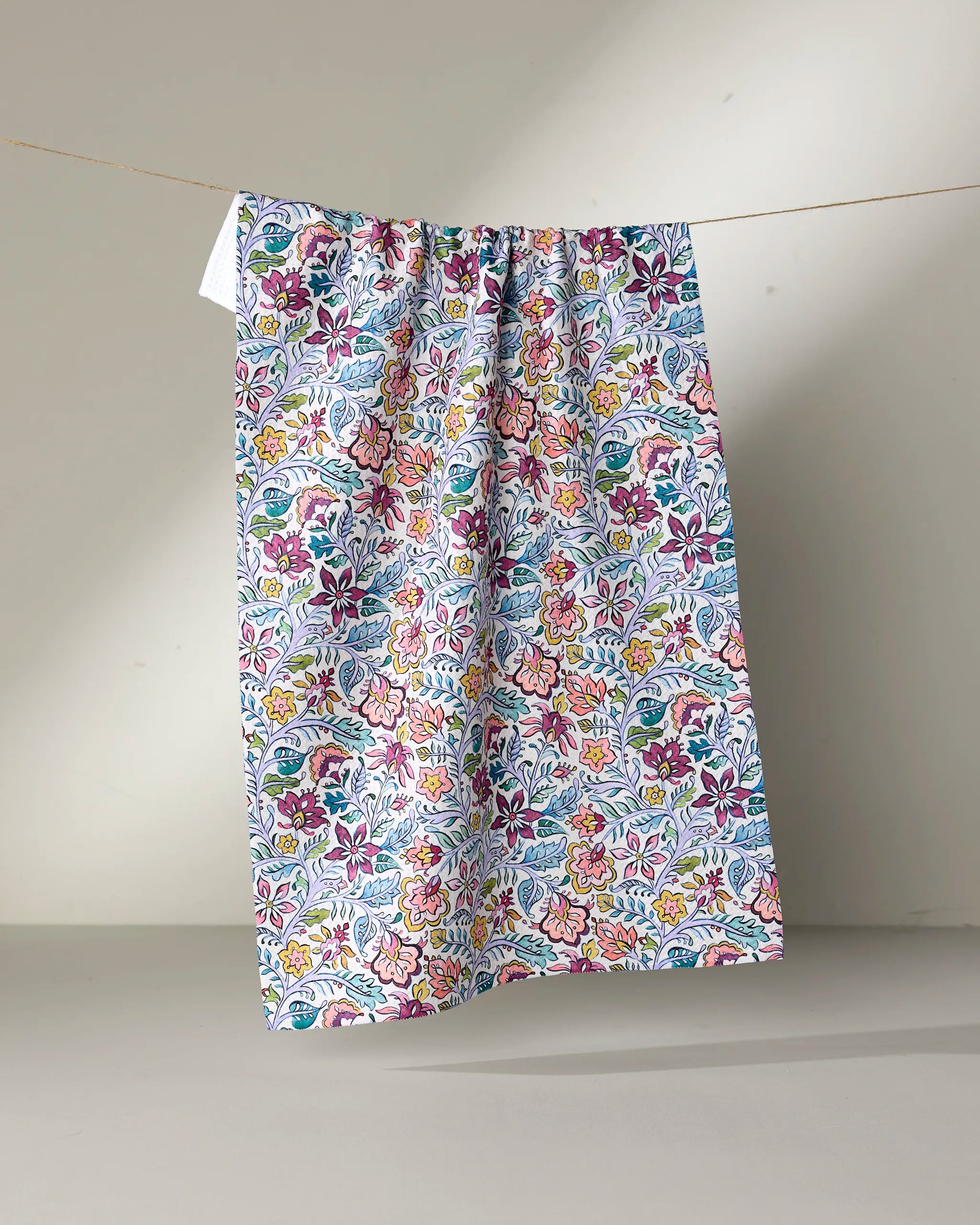 The Amethyst kitchen towel by Maayan Alper-Swan hangs from a string, showcasing its eco-friendly design and vibrant floral pattern in purple, pink, yellow, and blue against a neutral background.