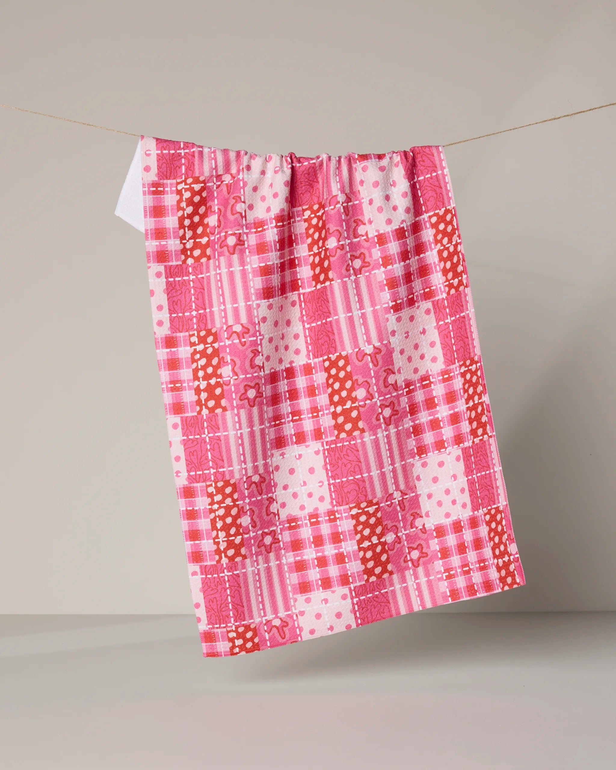 The Amor Amor towel by Geometry, featuring a pink patchwork pattern in various shades and designs, hangs ultra-absorbent on a clothesline against a neutral background.