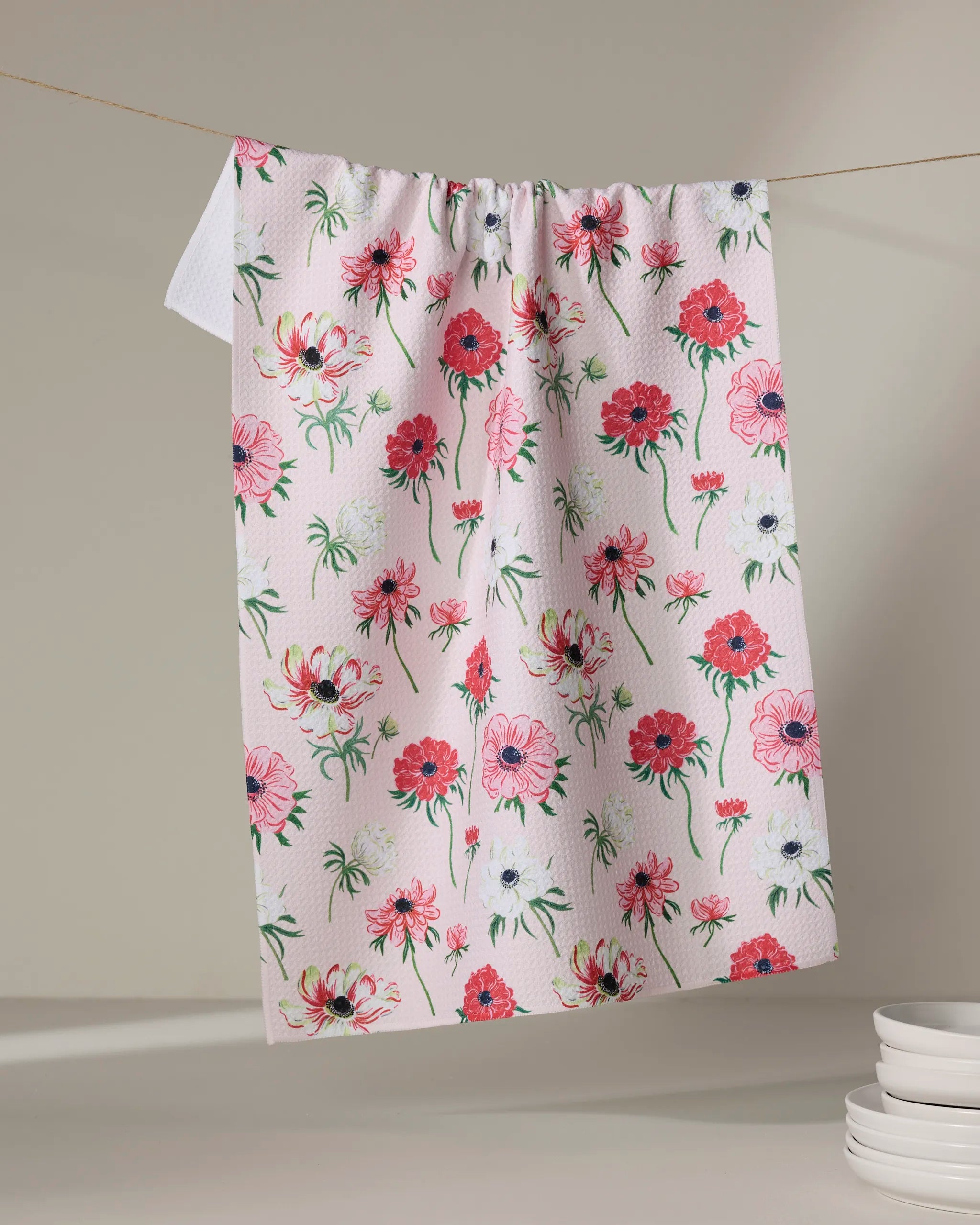 The Linda Fox Anemone Garden Flowers pink tea towel, featuring a colorful floral pattern, hangs on a clothesline with white plates nearby on a neutral surface.