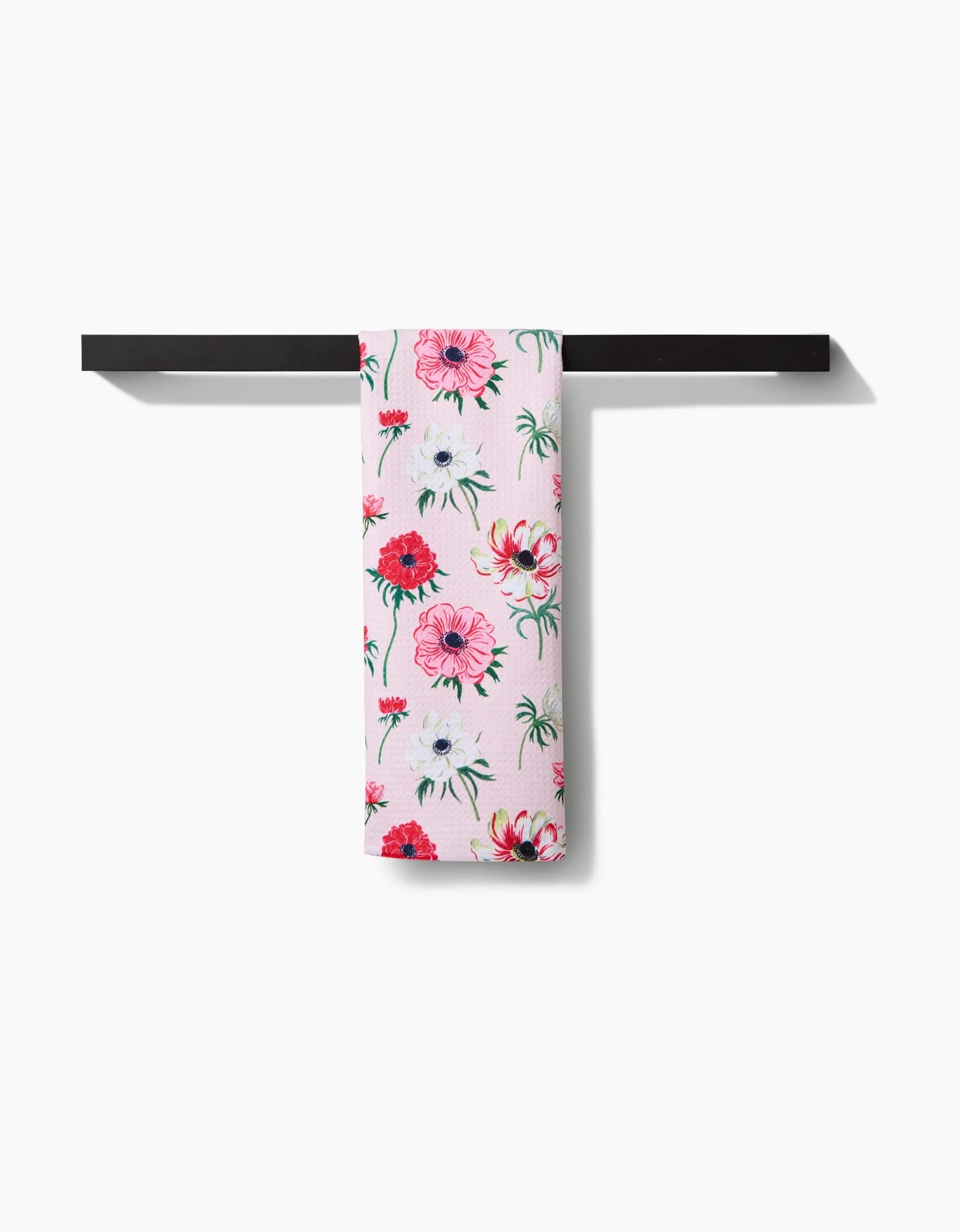 The Linda Fox Anemone Garden Flowers tea towel, featuring a pink floral pattern in red, white, and pink on durable Geoweave fabric, hangs neatly on a black bar against a white background.