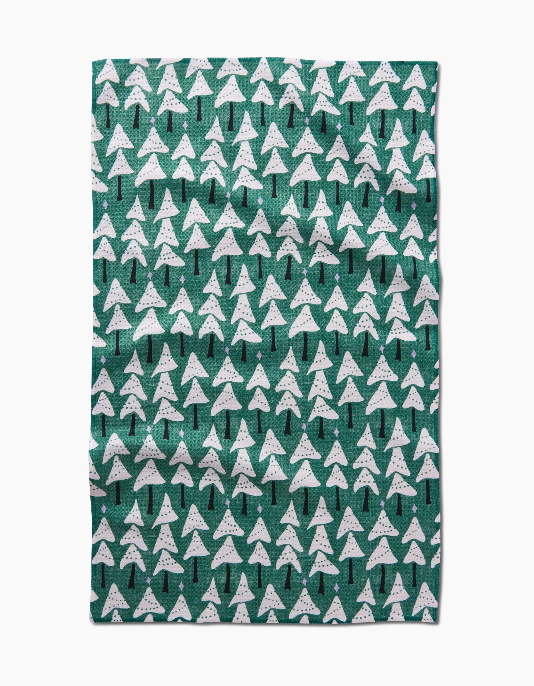 The Angelina by Amy MacCready is a green rectangular tea towel with white stylized pine trees and black trunks in rows across its ultra-absorbent surface. The fabric has a slightly wrinkled look.