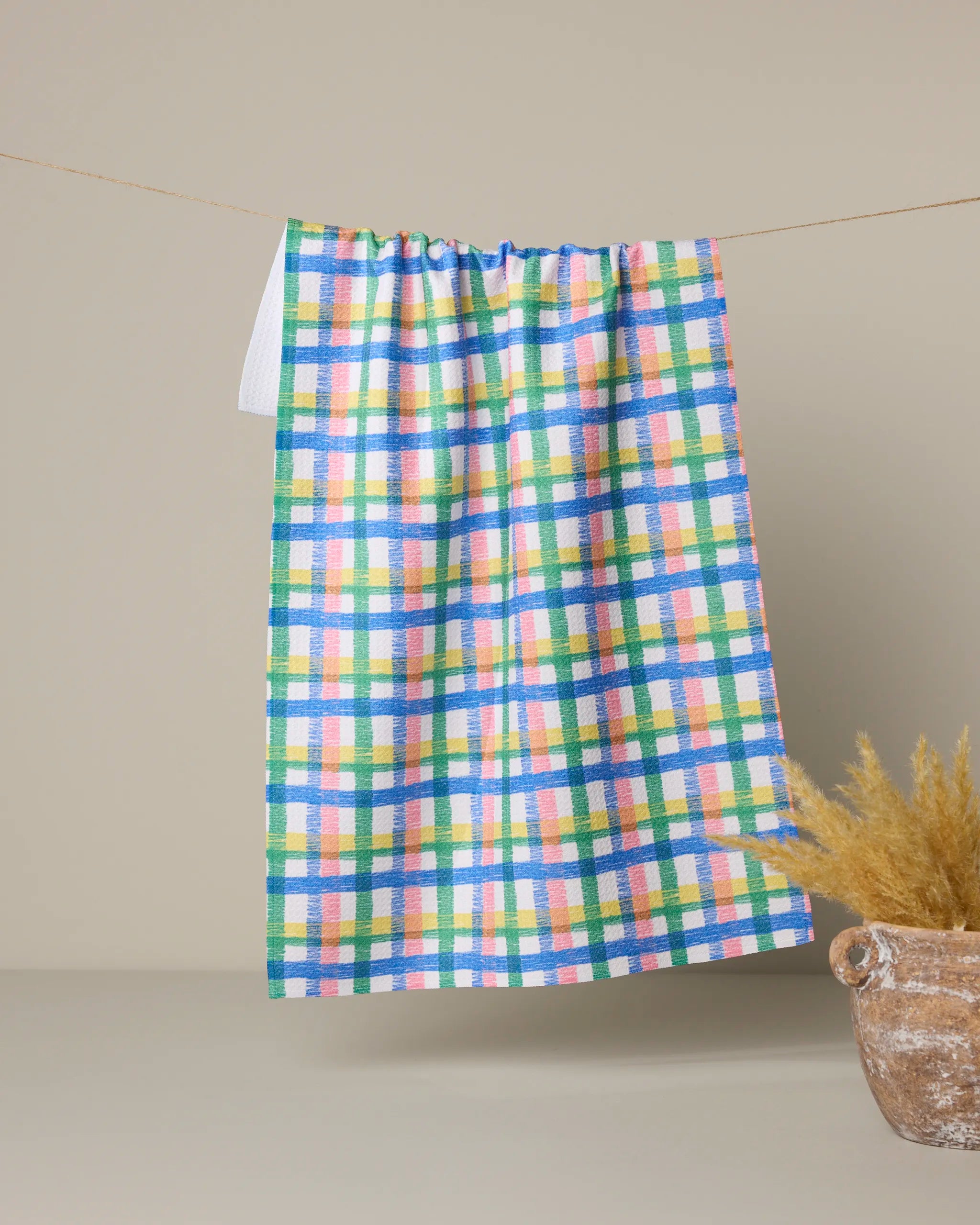 The Geometry Ansel tea towel, made from Geoweave fabric and featuring blue, green, yellow, pink, and white checks, hangs on a line against a beige background beside a rustic pot filled with dried grass.
