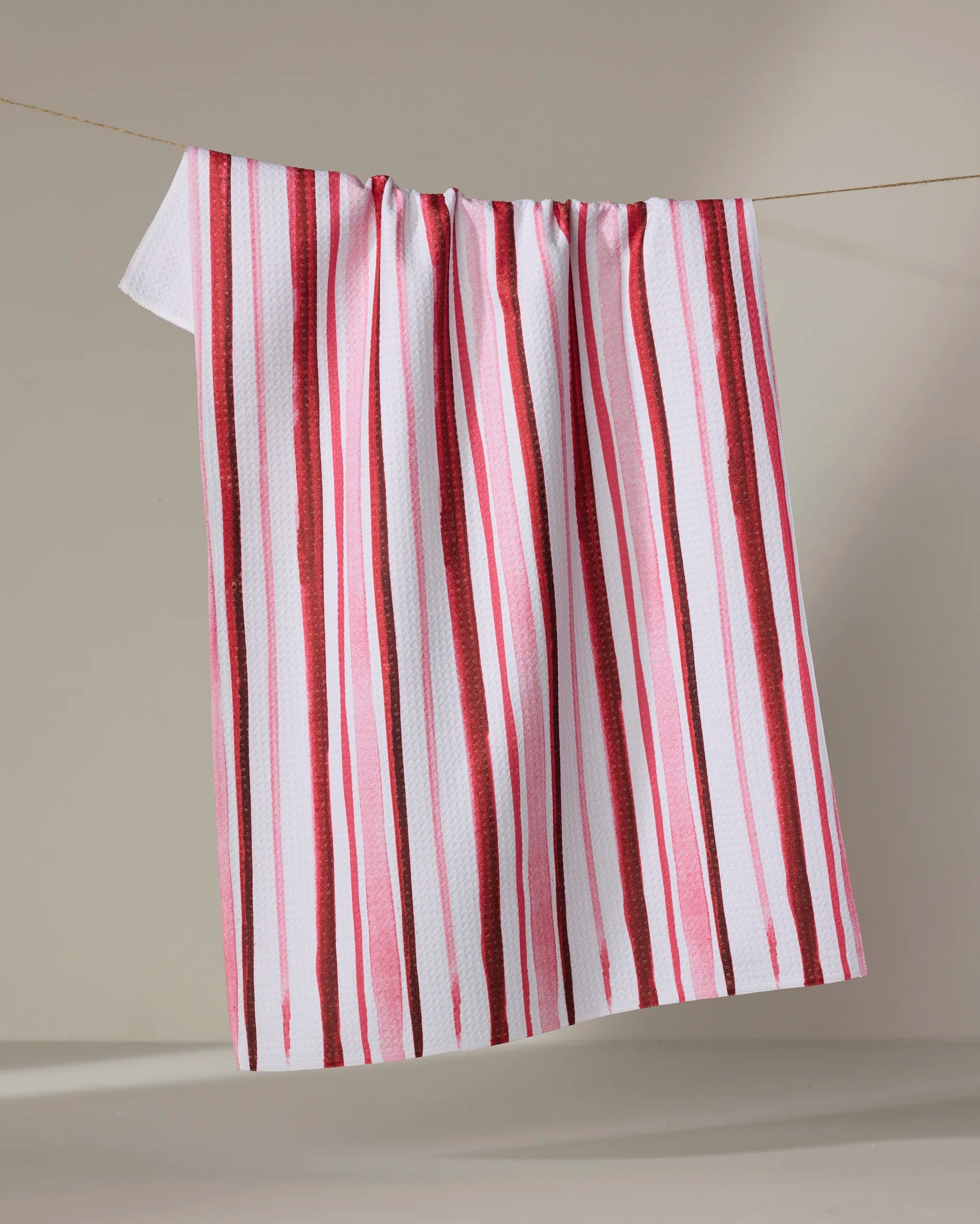 The Aperol in Rome Stripe kitchen towel by Travel Write Draw by Meagan Morrison, featuring recycled fabric and vertical red and pink stripes, hangs on a clothesline against a neutral background.